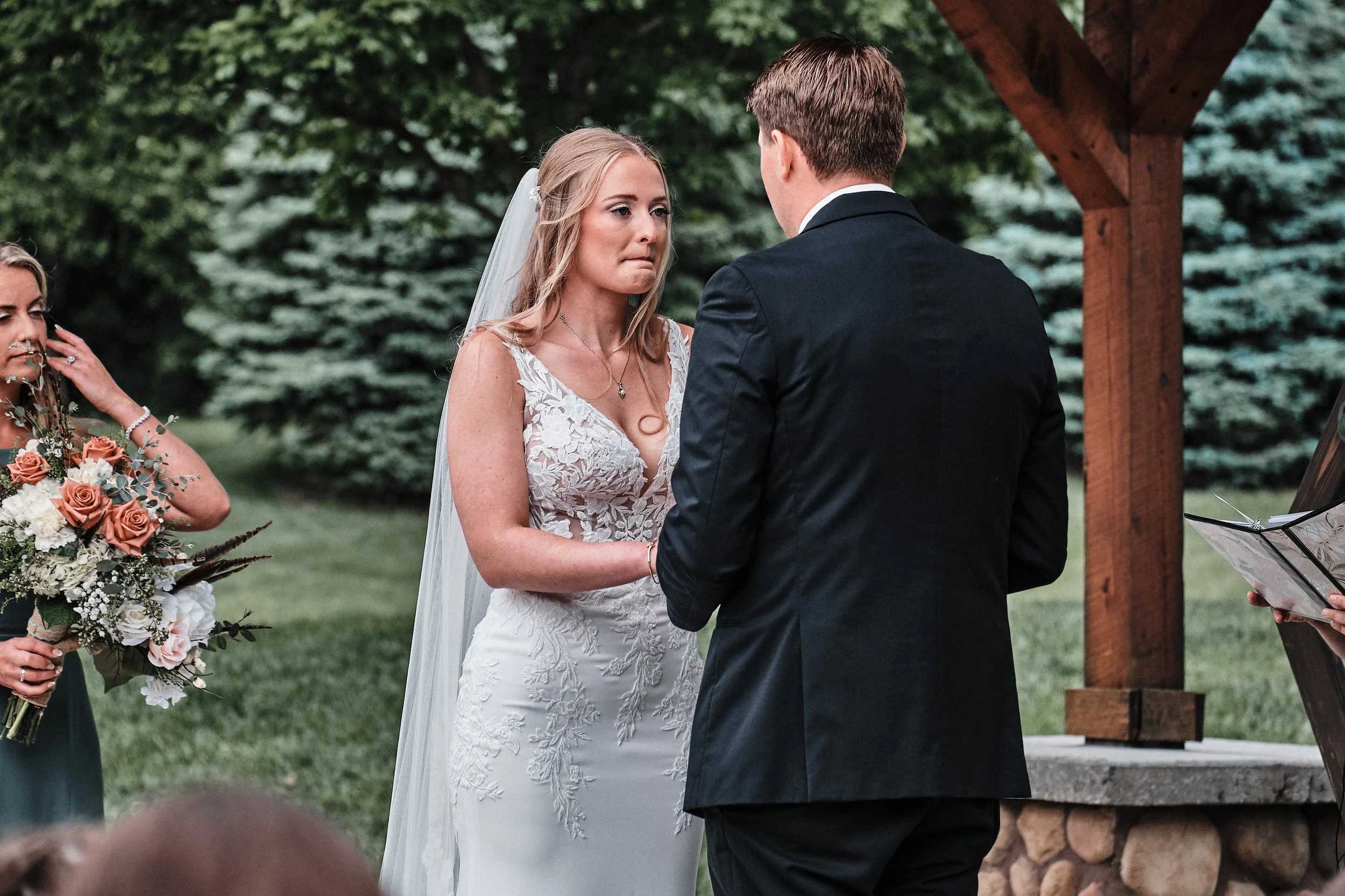A bride and groom exchange vows during their outdoor wedding ceremony, with a bridesmaid holding a bouquet of flowers nearby.