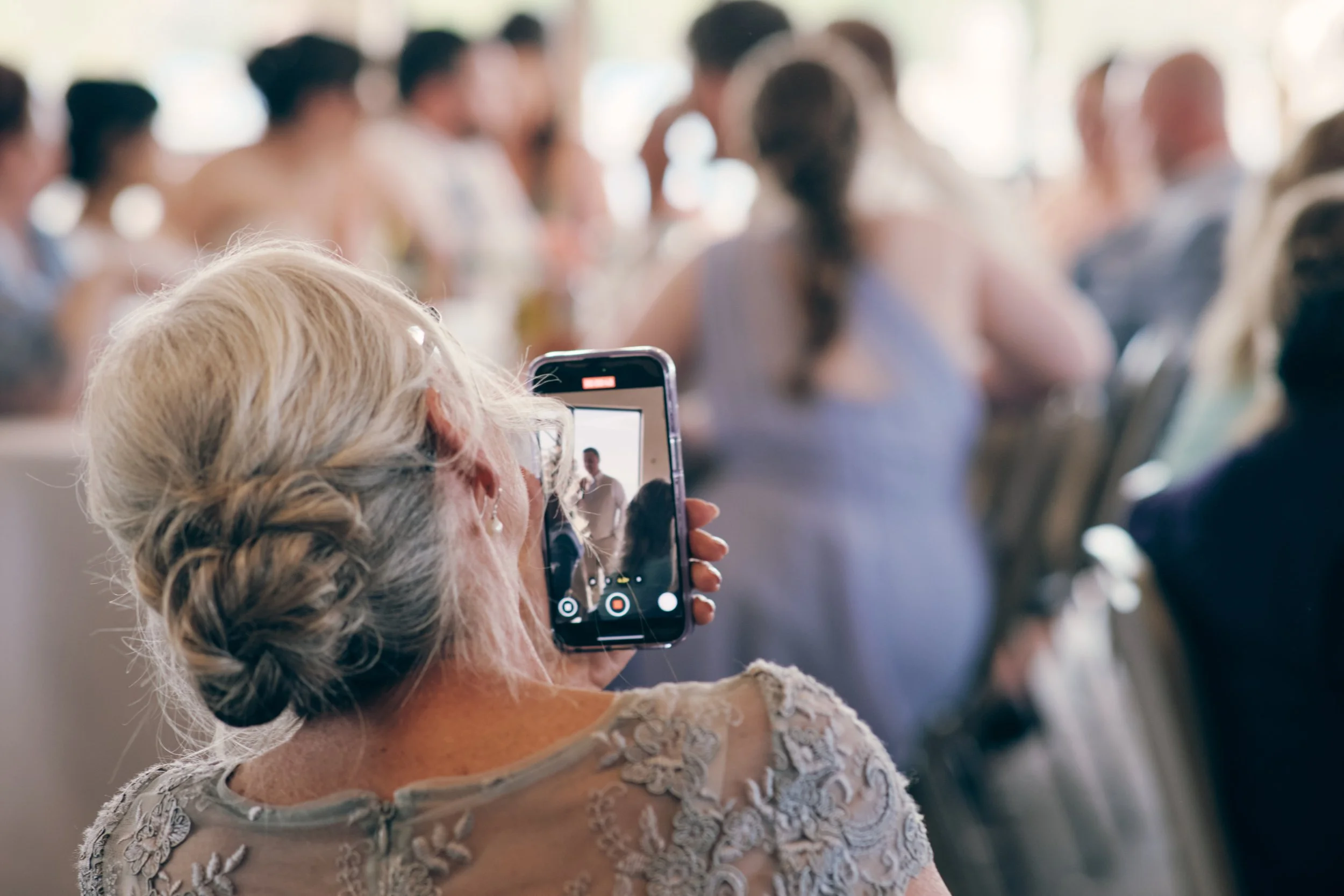An elderly woman with white hair styled in a braid, wearing a lace dress, is taking a photo or video with her smartphone at an indoor social gathering. Other people are sitting in the background, blurred, suggesting a crowd or event.