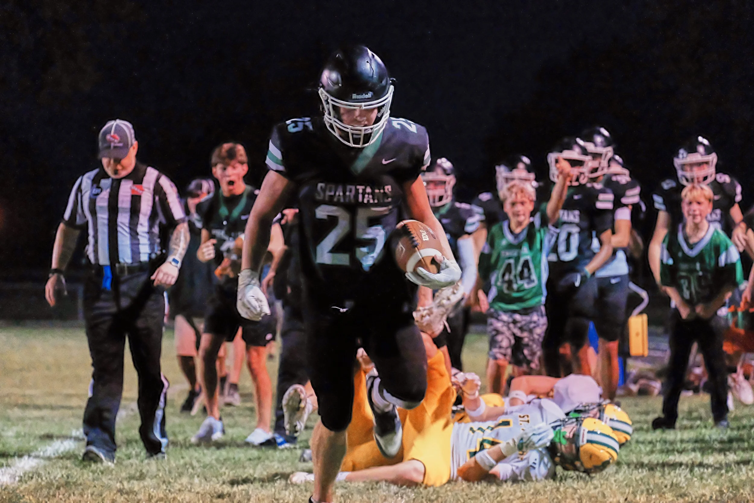 A youth football player in black jersey holding a football, running past a fallen opponent in yellow jersey, with a group of cheering children and referee in the background on a football field at night.