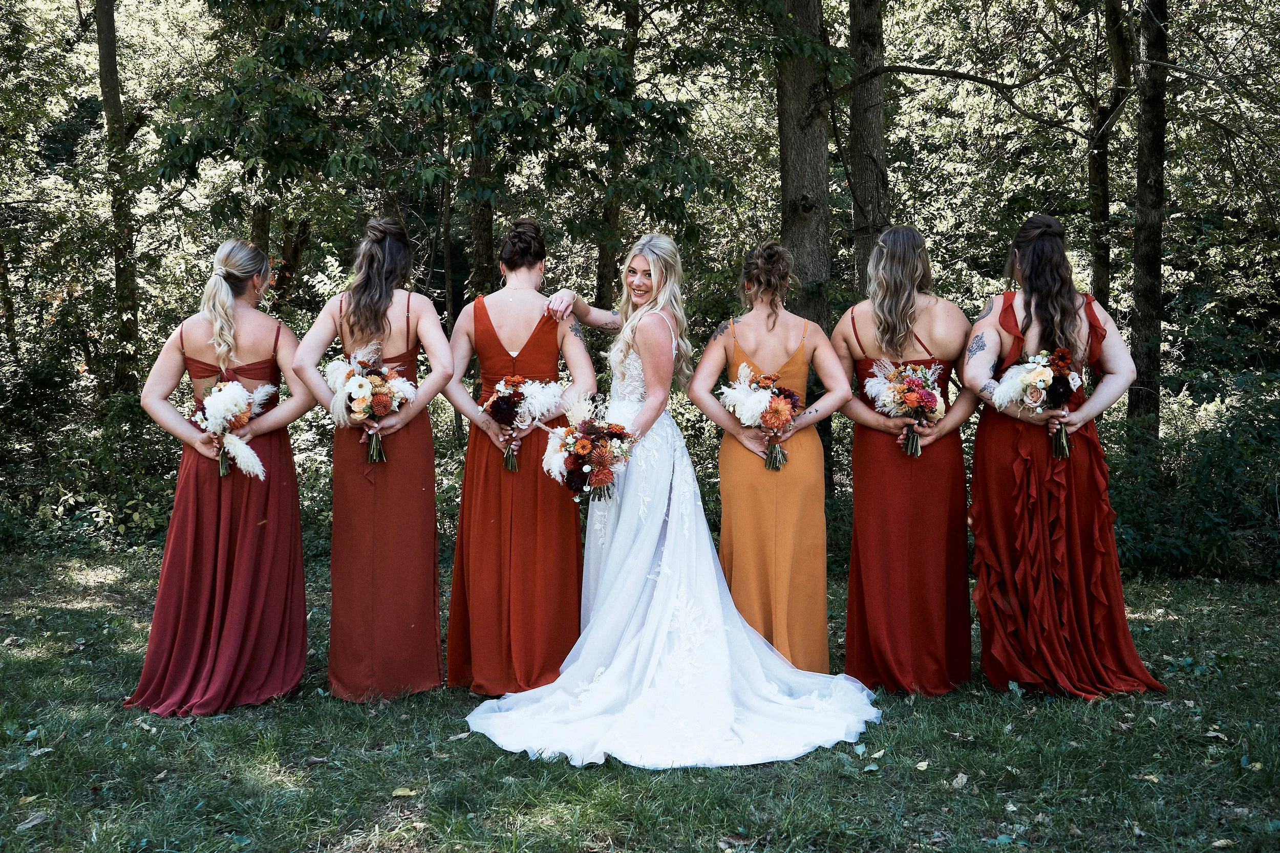 Group of women in bridesmaid dresses standing outdoors among trees, holding bouquets, with one woman in a white wedding dress smiling at the camera.
