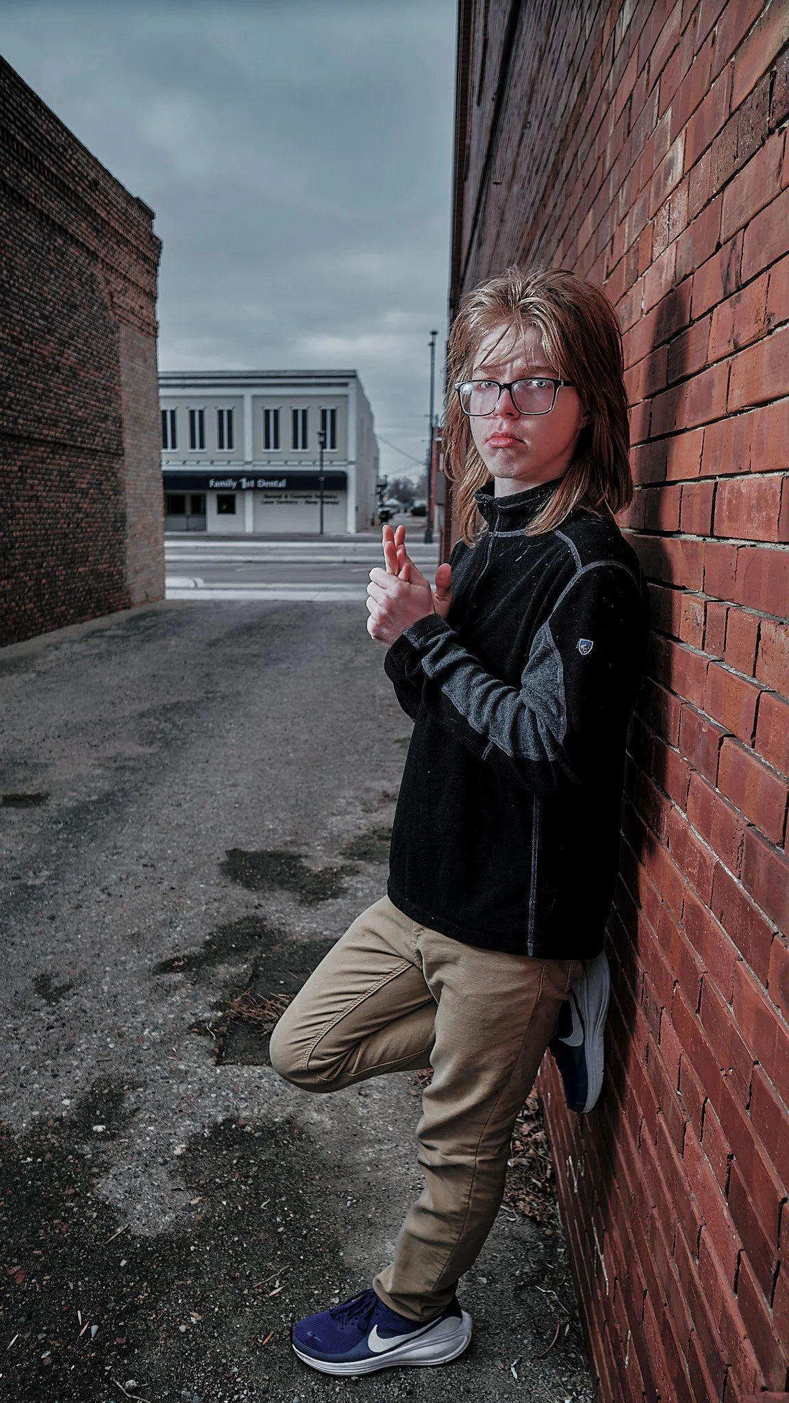 A young man with glasses and long hair leaning against a brick wall outdoors, with crossed hands and one leg bent, wearing a black jacket, khaki pants, and blue sneakers, in an urban setting with a cloudy sky and buildings in the background.