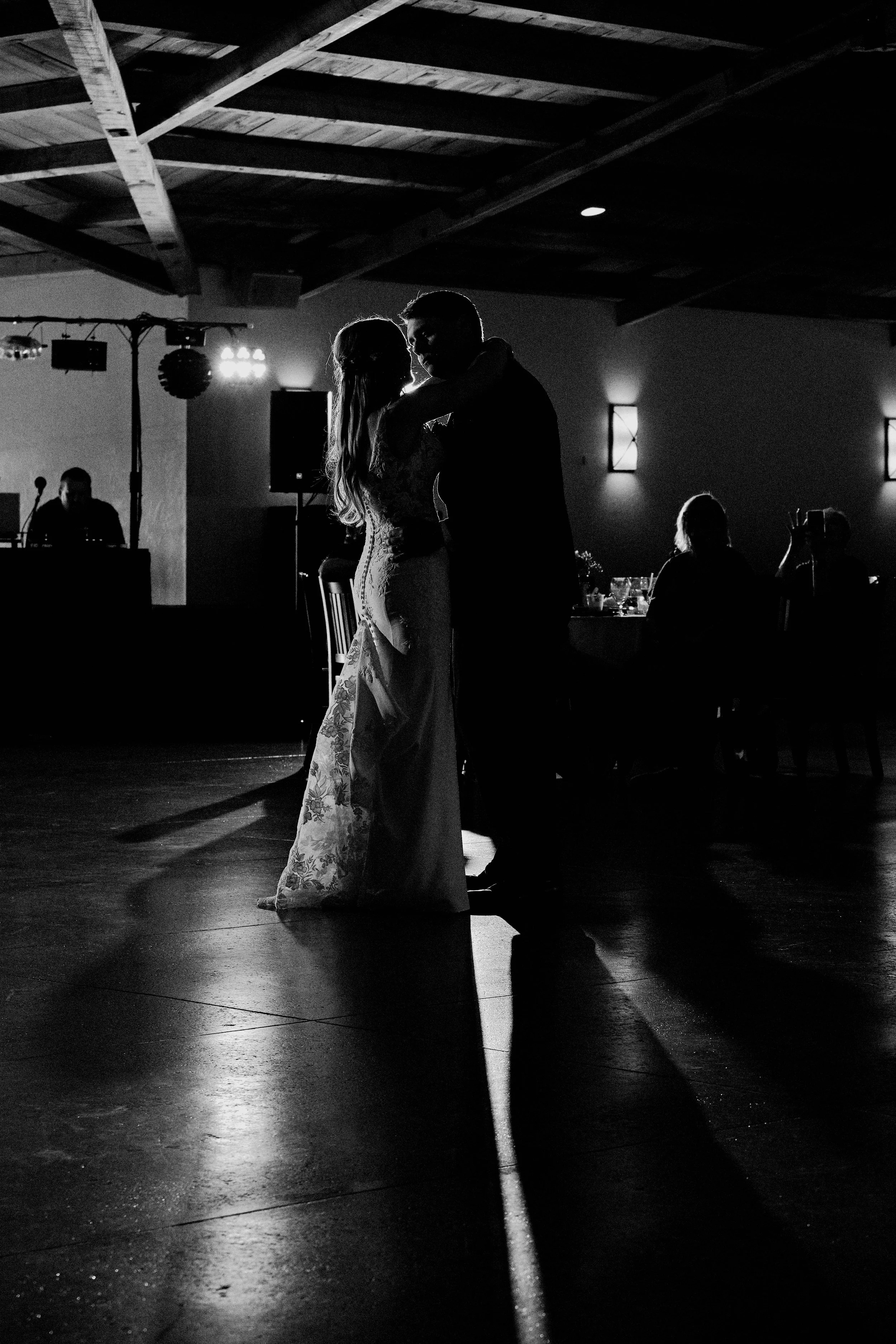 Silhouette of a couple dancing in a dimly lit indoor venue, with a wooden ceiling and some seated guests visible in the background.