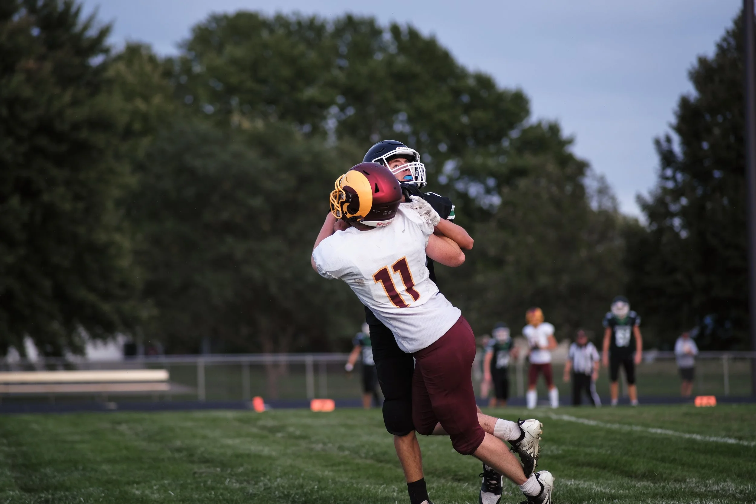 A football player in a white uniform with the number 11 is celebrating a successful play, lifting a teammate with a maroon and yellow helmet on a football field during a game at sunset.