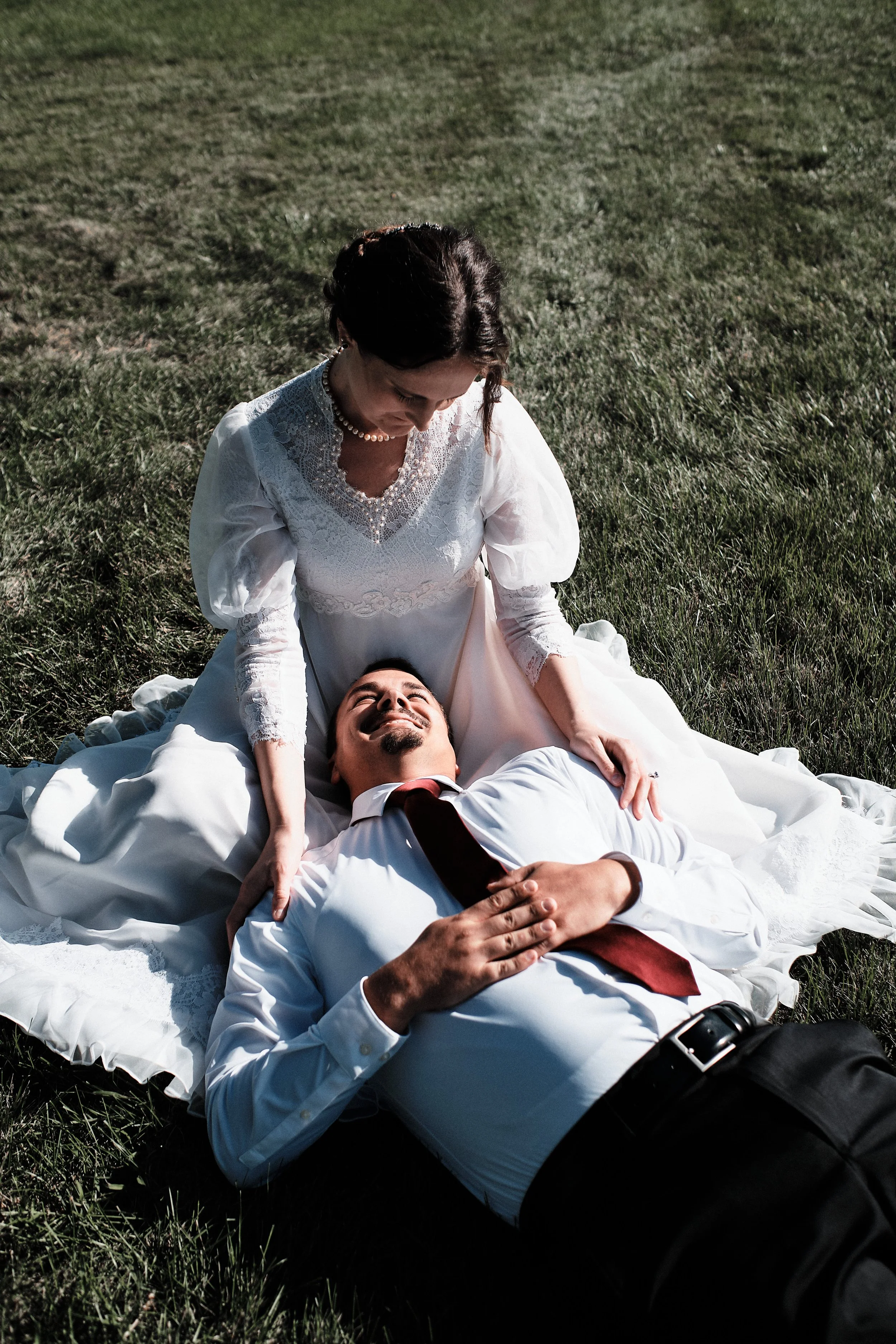 A man and woman in wedding attire lying on the grass outdoors; the woman is seated over the man, looking down at him with a gentle expression, while the man is lying on his back, smiling, with his hand on his chest.