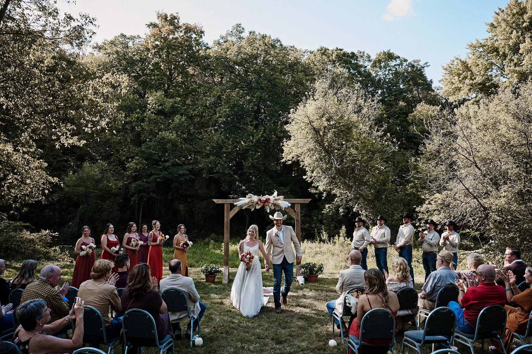 A wedding ceremony outdoors in a wooded area, with the bride and groom walking hand-in-hand down an aisle. Groups of bridesmaids and groomsmen stand on either side, and seated guests watch the event.
