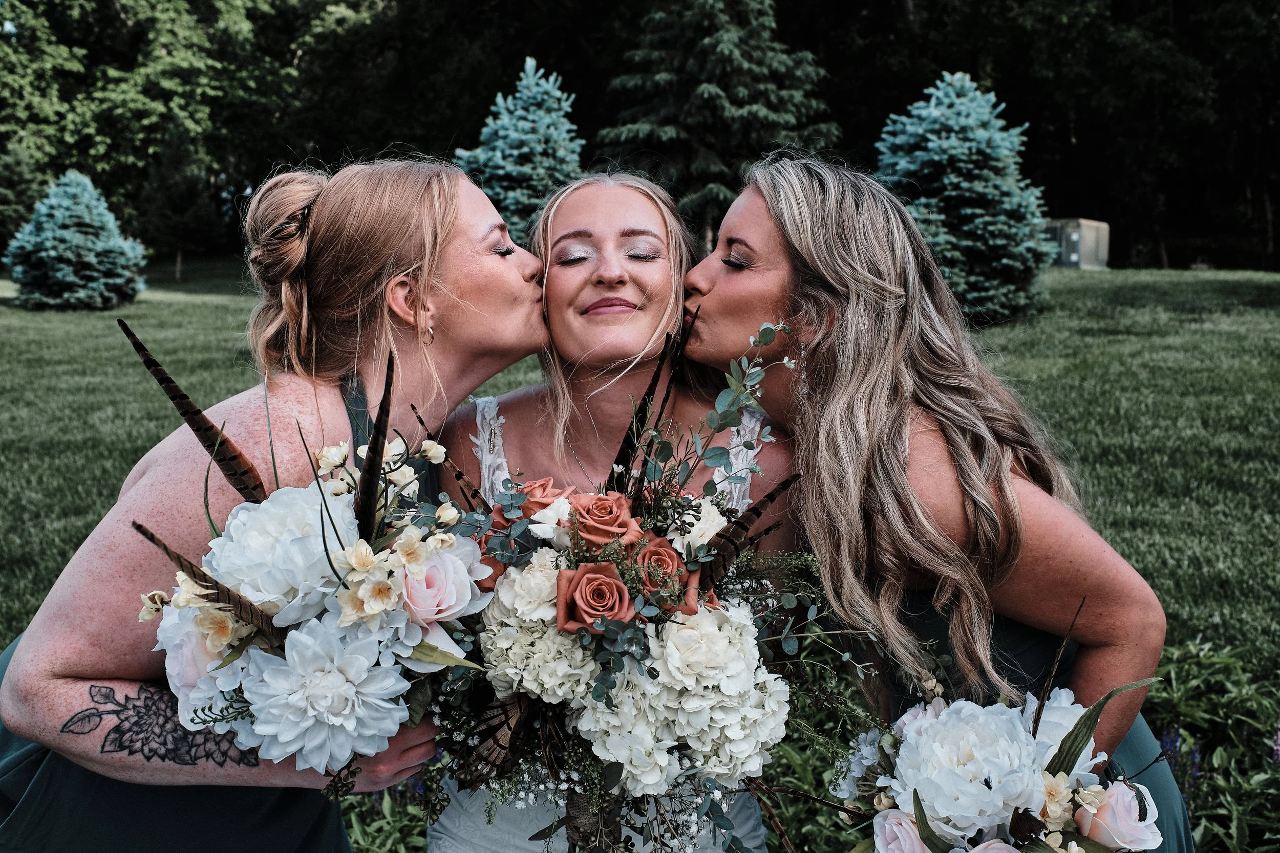 Three women, two with blonde hair and one with brown hair, are kissing a woman with blonde hair on each cheek at a wedding or celebration outdoors, with evergreen trees and a grassy area in the background. The women are holding a large bouquet of flo