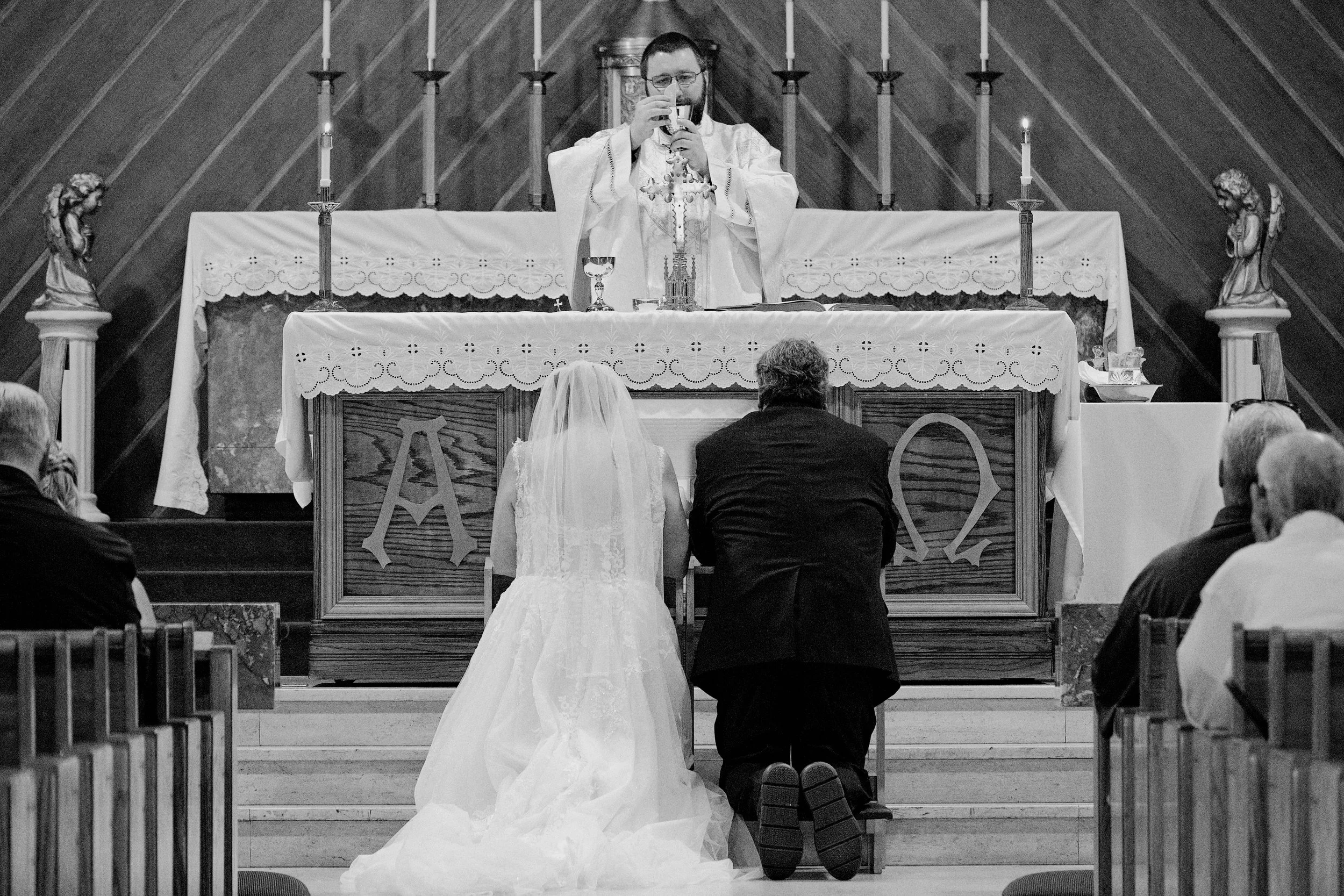 A priest in religious vestments holding a chalice during a wedding ceremony in a church, facing a bride and groom kneeling at the altar.