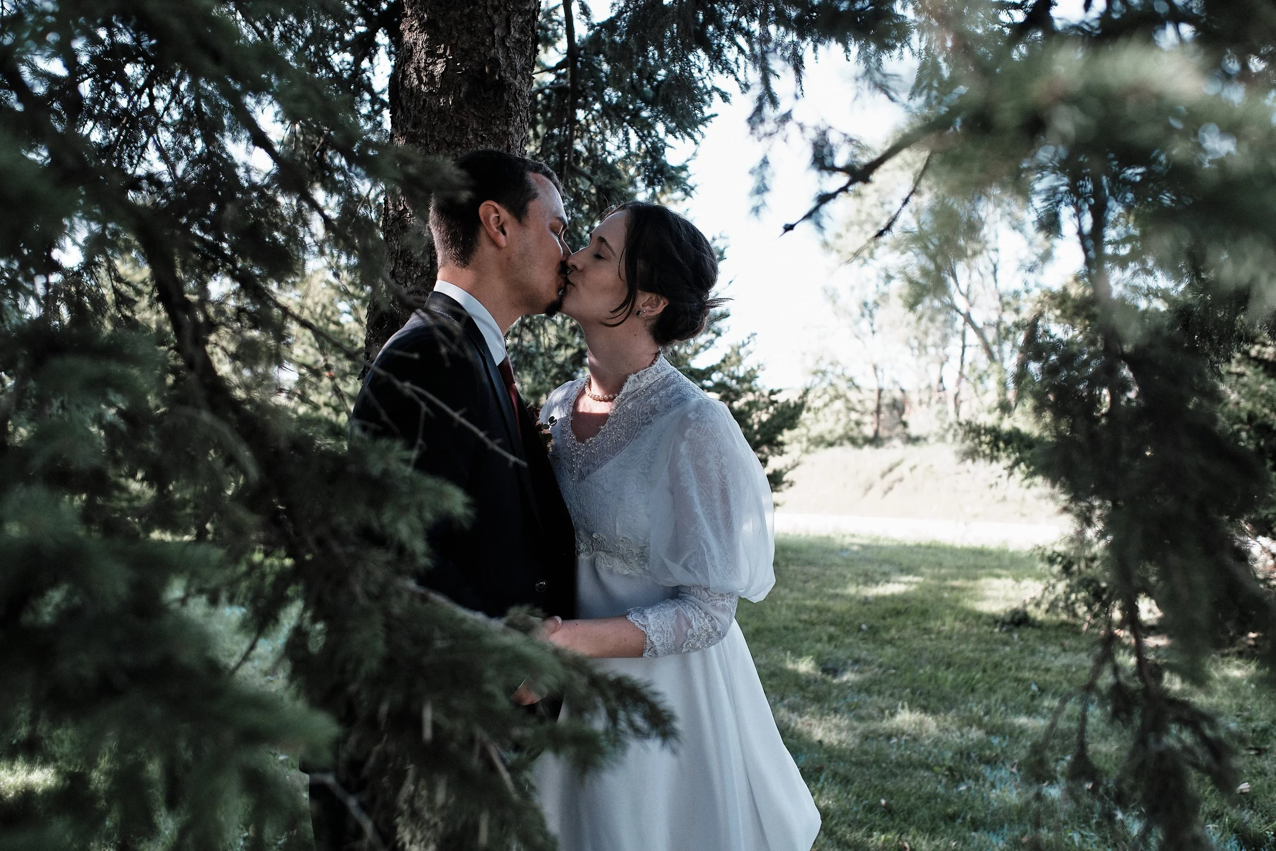 A couple, dressed in wedding attire, sharing a kiss outdoors among trees.