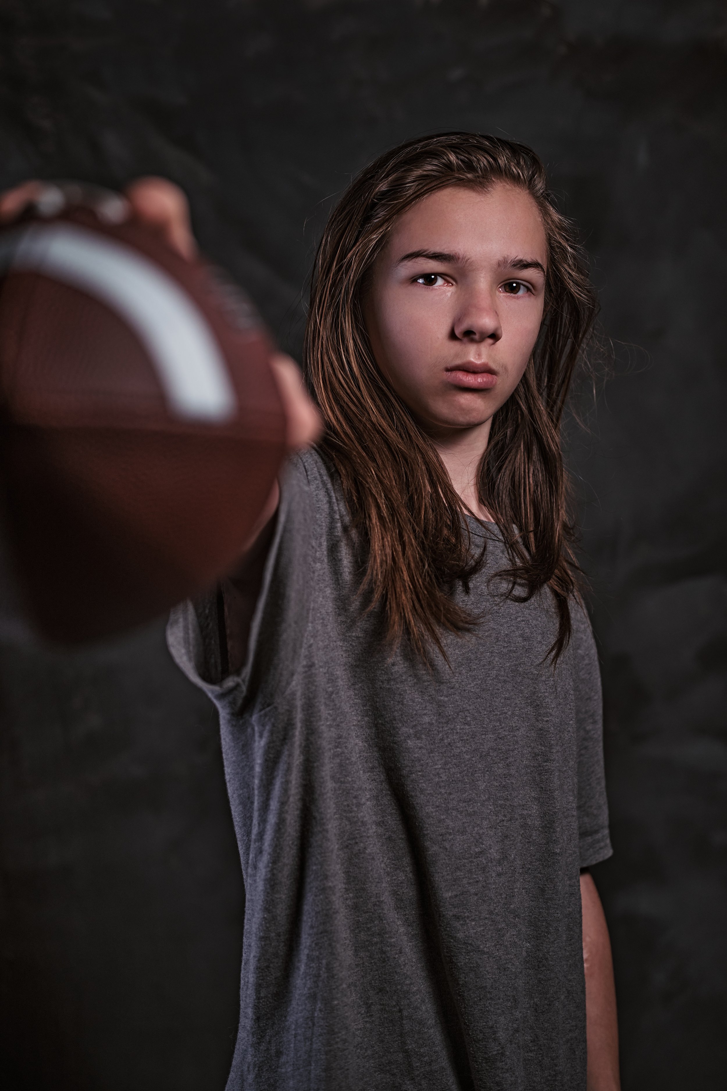 A young person with long brown hair, wearing a gray t-shirt, holding a football towards the camera, with a serious expression against a dark background.