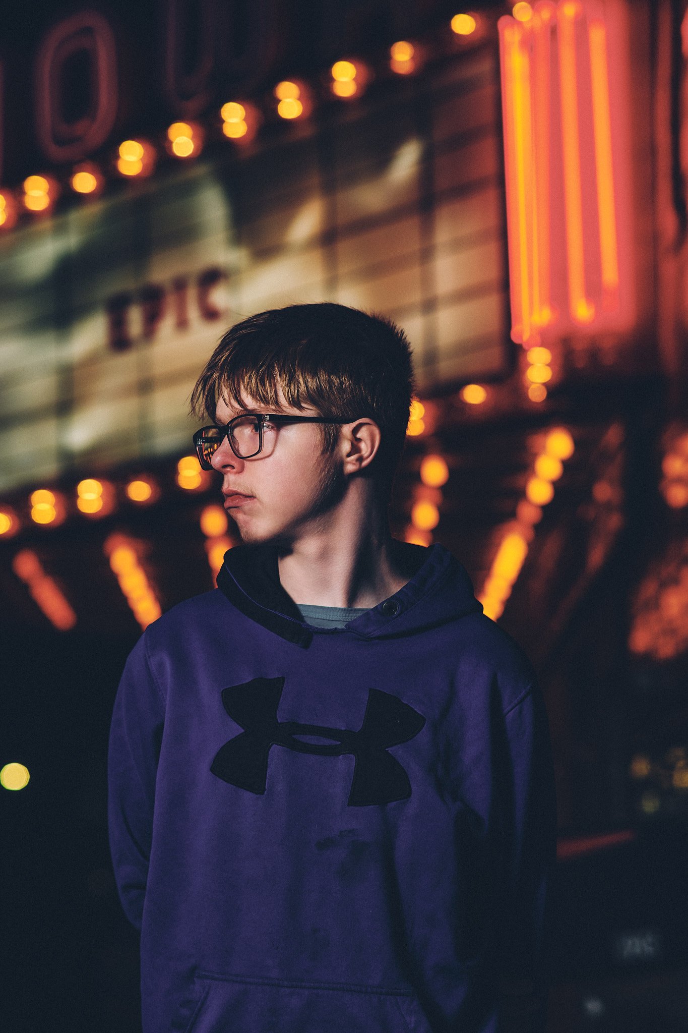 A young person with short, dark hair and glasses standing in front of a neon-lit background, wearing a purple Under Armour hoodie.