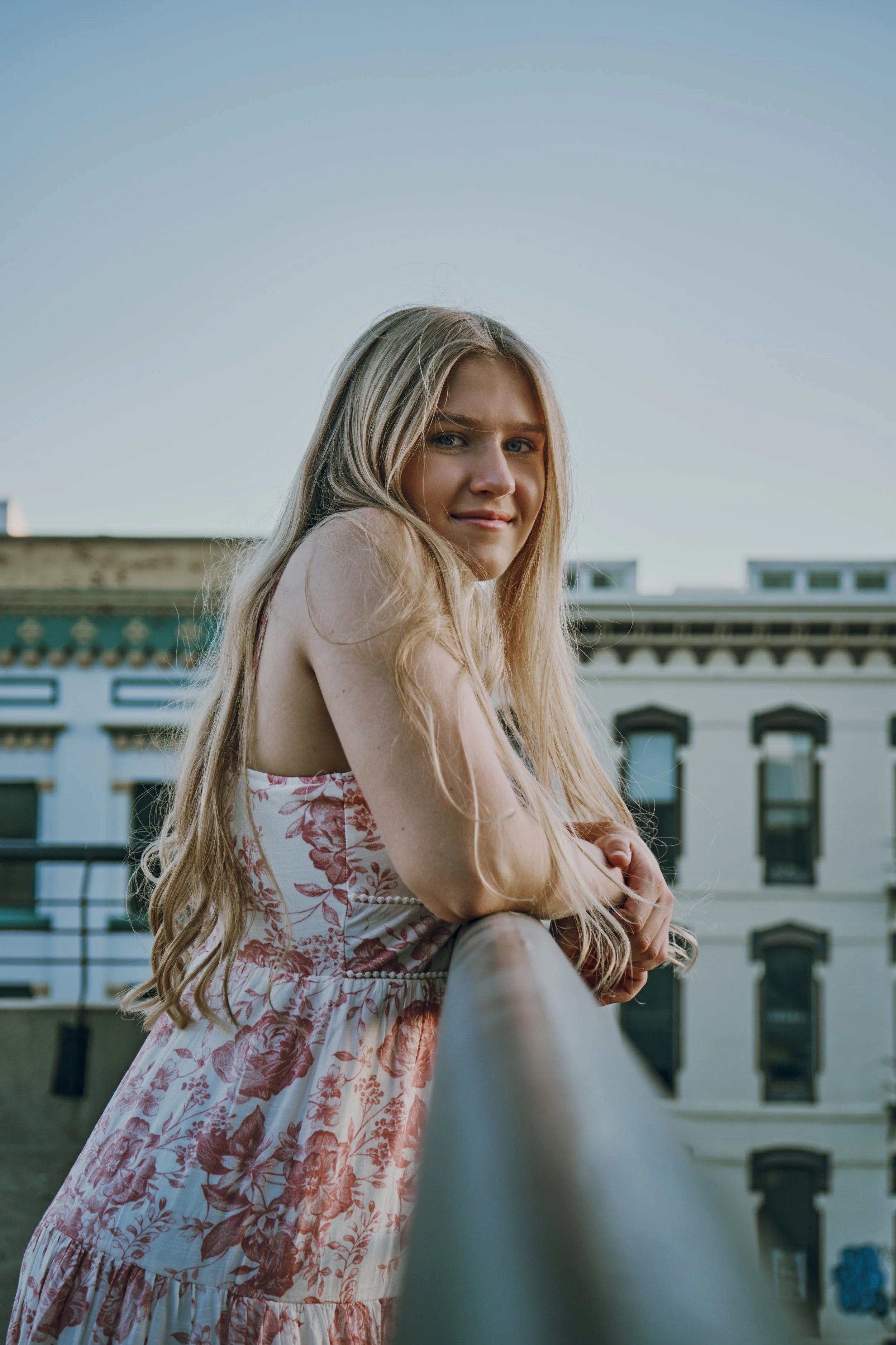 A young woman with long blonde hair sitting outdoors on a ledge, looking over her shoulder, with a city building in the background during the daytime.