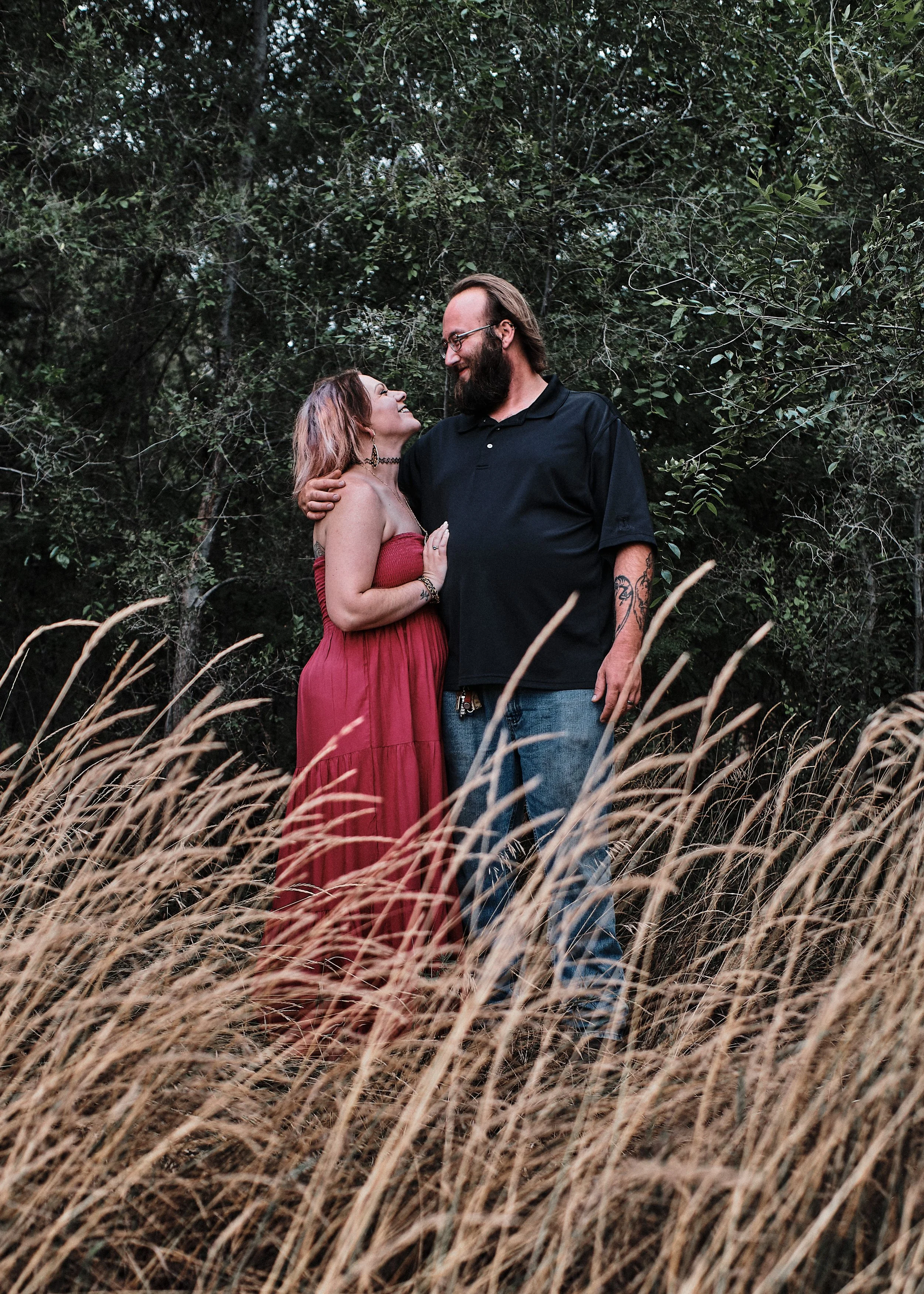A couple standing close together outdoors in a natural setting with trees and grass, looking lovingly at each other.