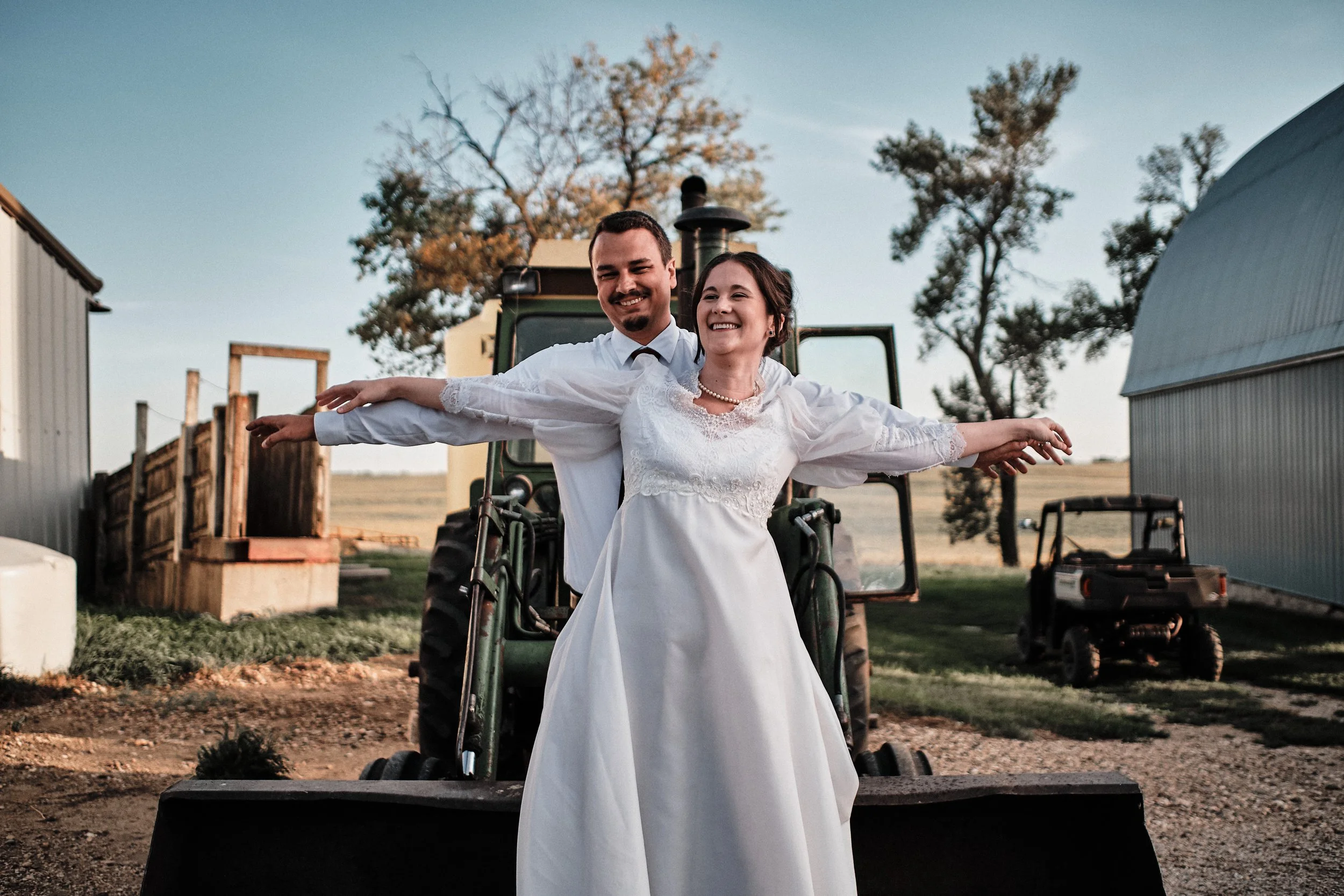 A newlywed couple standing on farm equipment, smiling, with trees and farm buildings in the background during sunset.