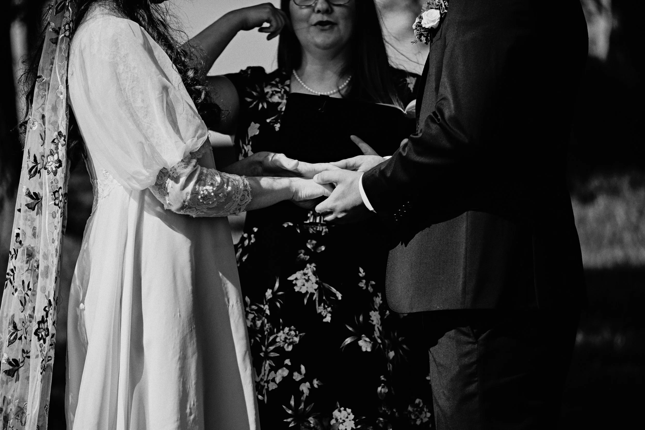 A black and white photo of a wedding ceremony showing a bride and groom holding hands, with an officiant reading from a book.