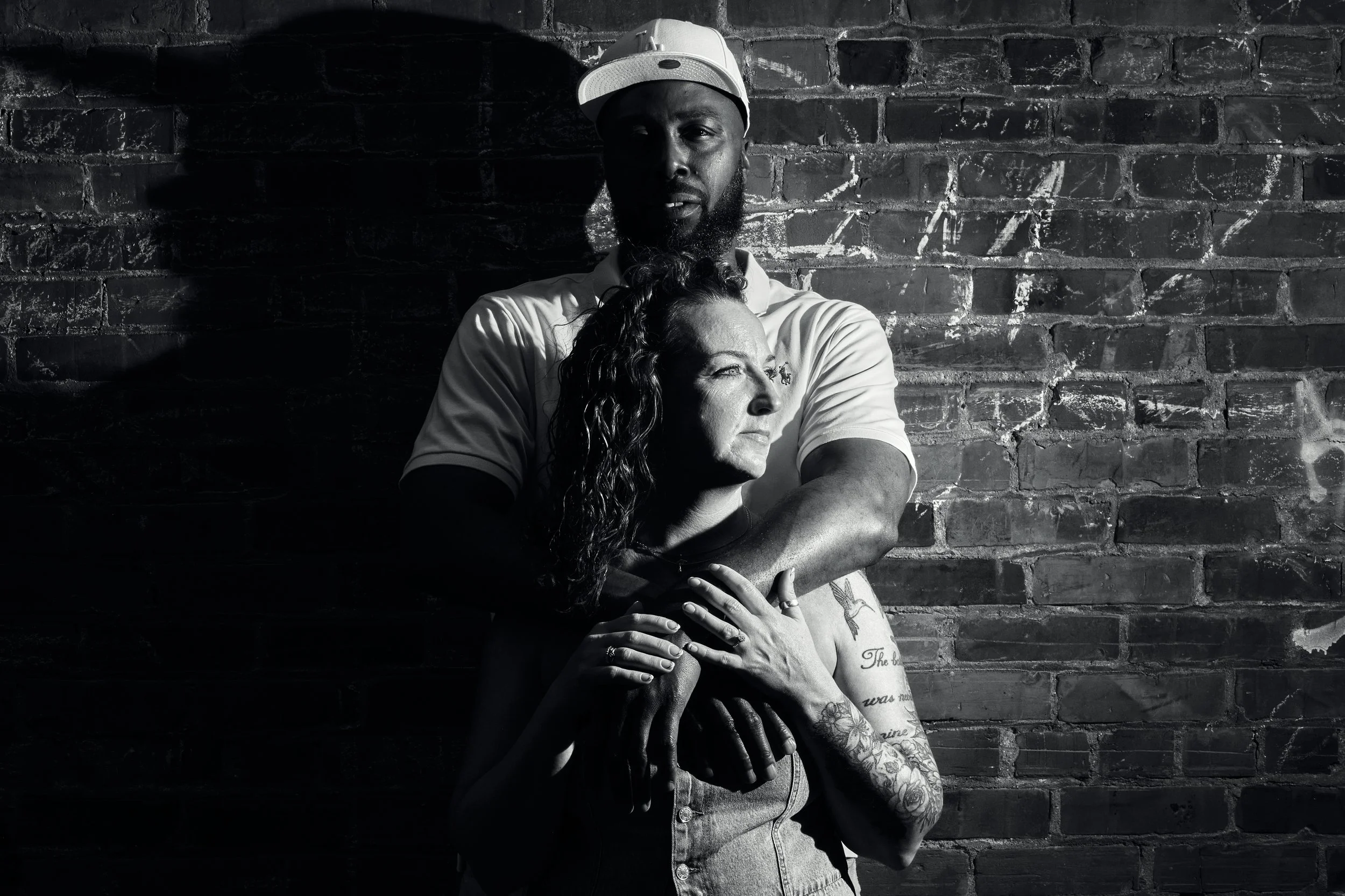 A black-and-white photo of a man wearing a baseball cap, standing against a brick wall. The man has a beard and is embracing a woman with curly hair and tattoos on her arm, who is resting her head on his chest. They are both looking in different dire