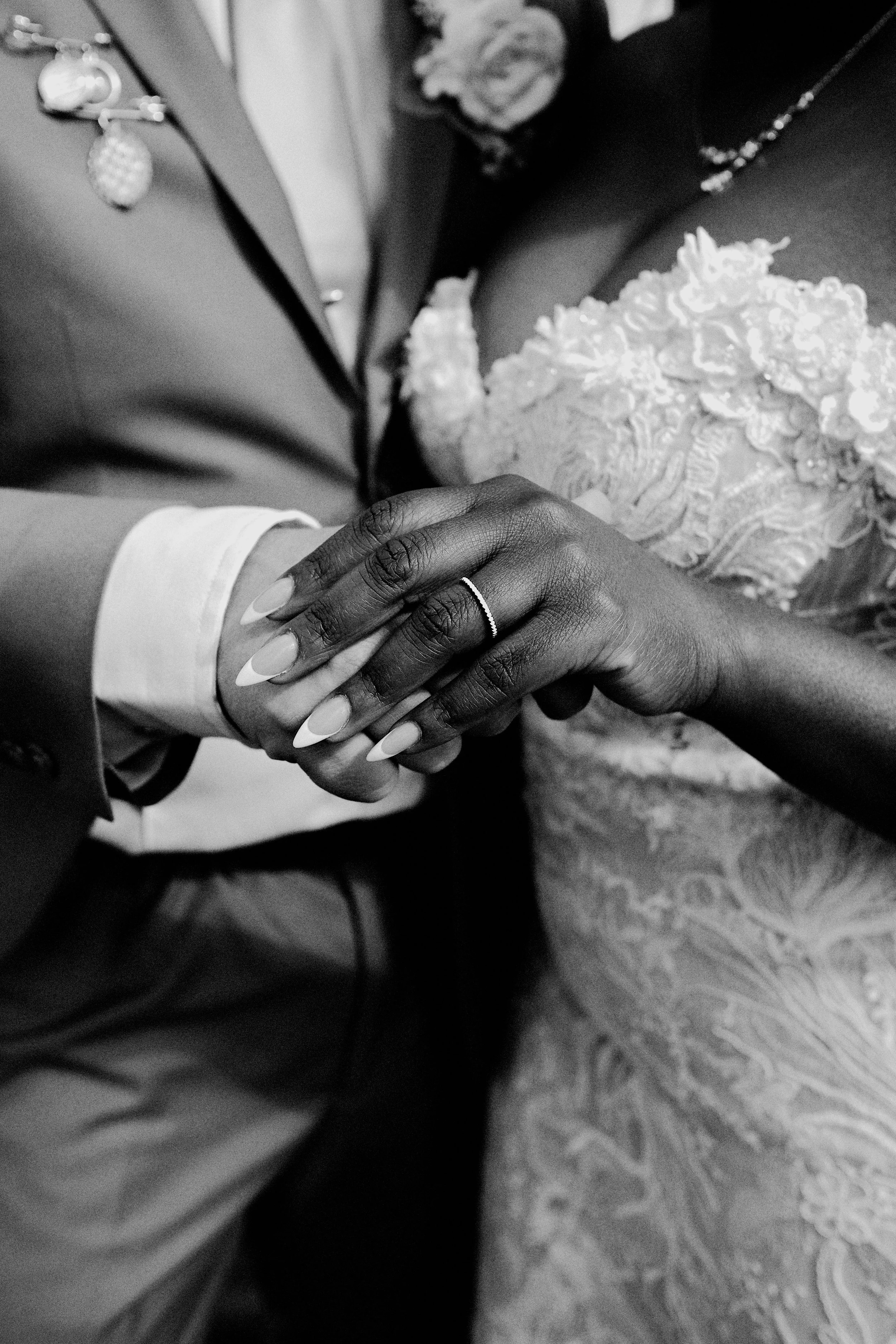 A close-up of a couple holding hands, with one hand being darker-skinned and the other lighter-skinned. The darker-skinned hand is wearing a ring. The background includes wedding clothing and jewelry.