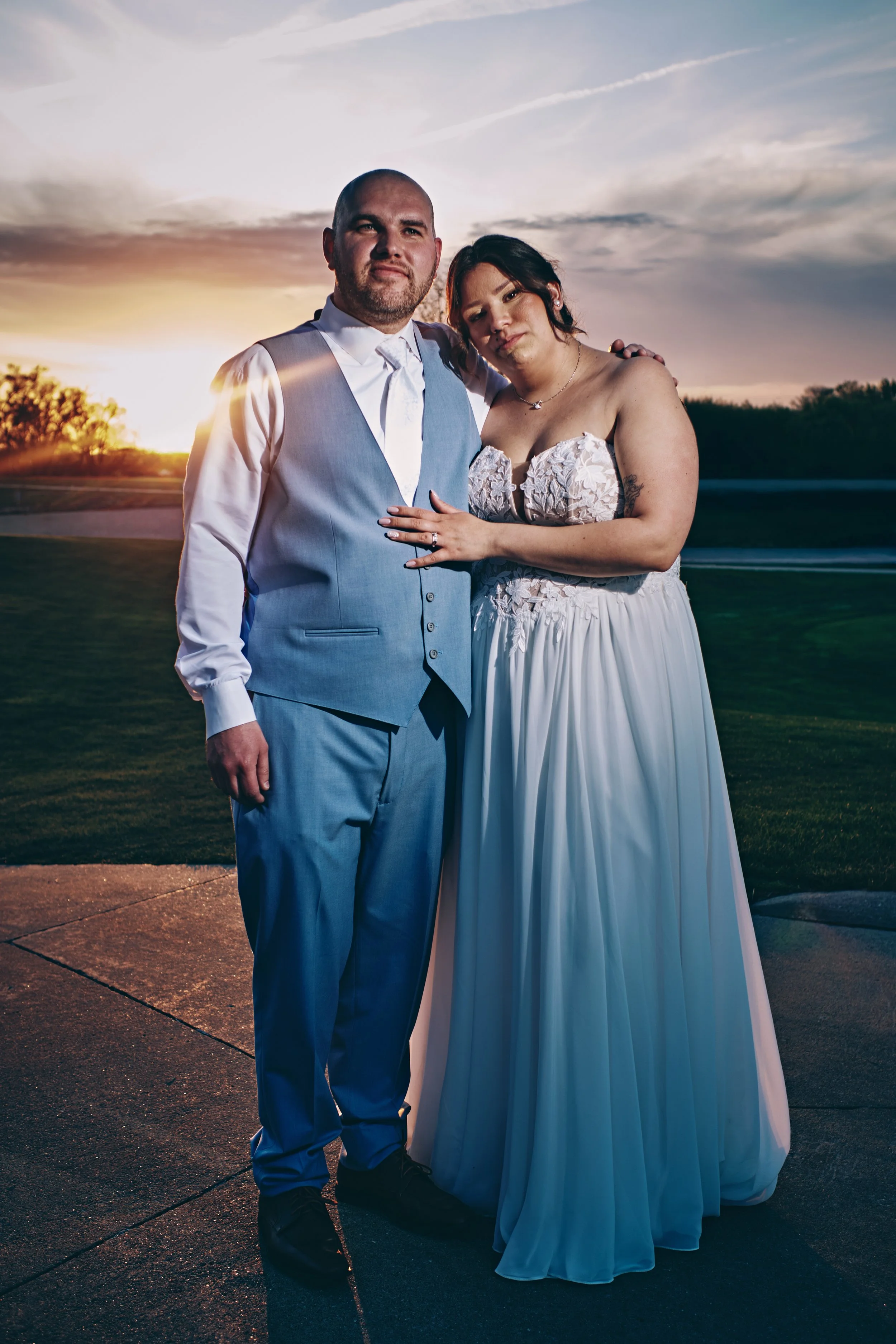 A man and woman in wedding attire standing outdoors during sunset, with the man in a light blue vest and pants, and the woman in a strapless white dress. They are close together, with the woman resting her hand on the man's chest.