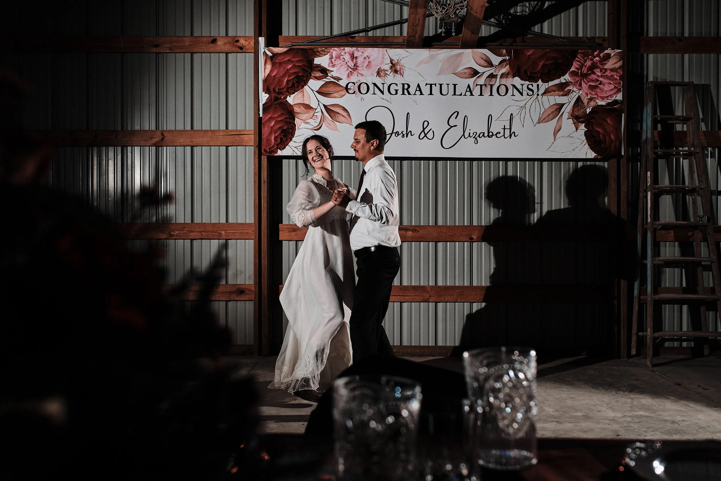 A bride and groom dancing at their wedding reception in a rustic indoor venue with a metal wall and wooden framework, a large floral banner reading 'Congratulations! Josh & Elizabeth,' and blurred guests in the foreground.
