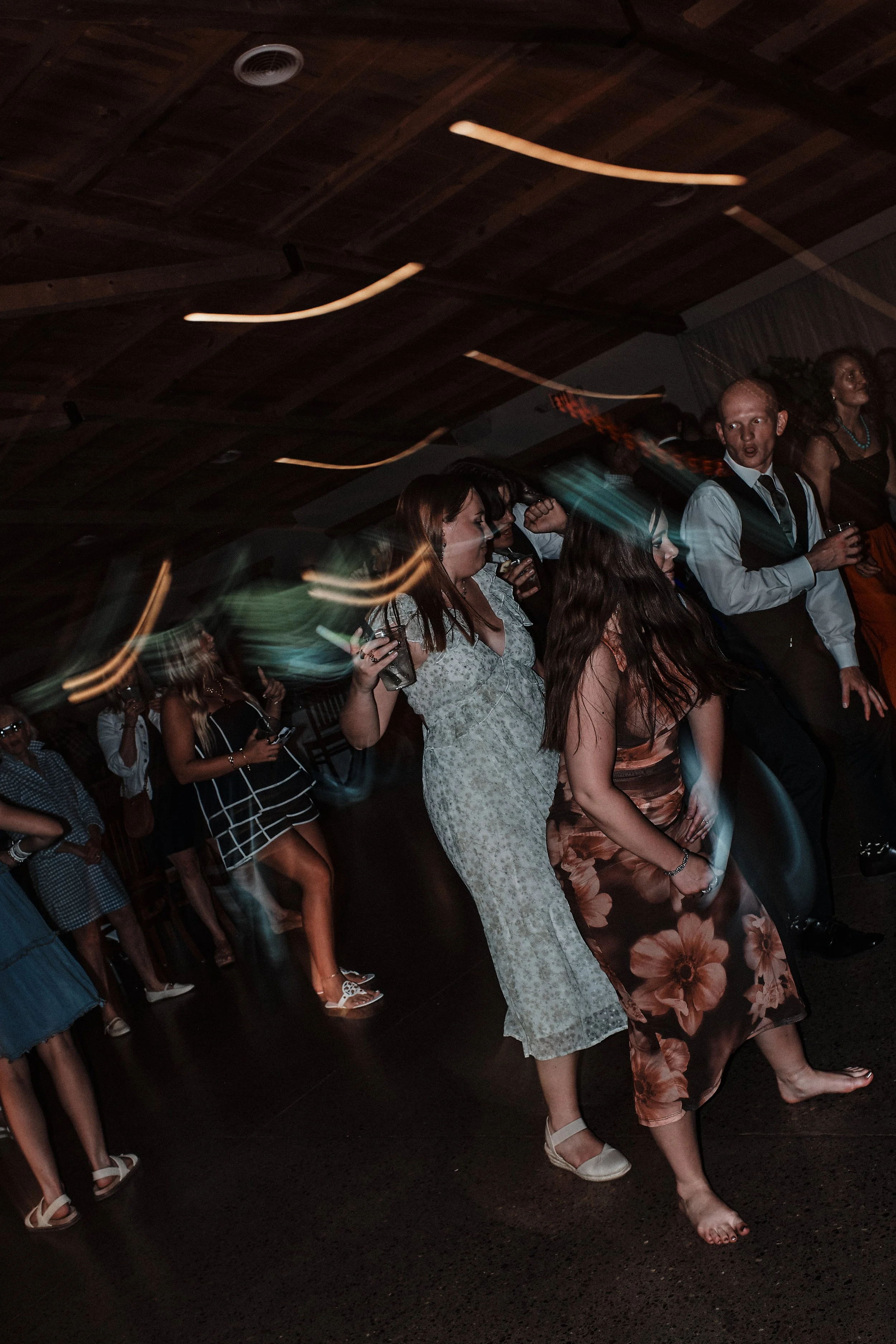 A group of people dancing at a party or social gathering in a dimly lit venue. The crowd includes both men and women, some are holding drinks, and one woman appears to be barefoot.