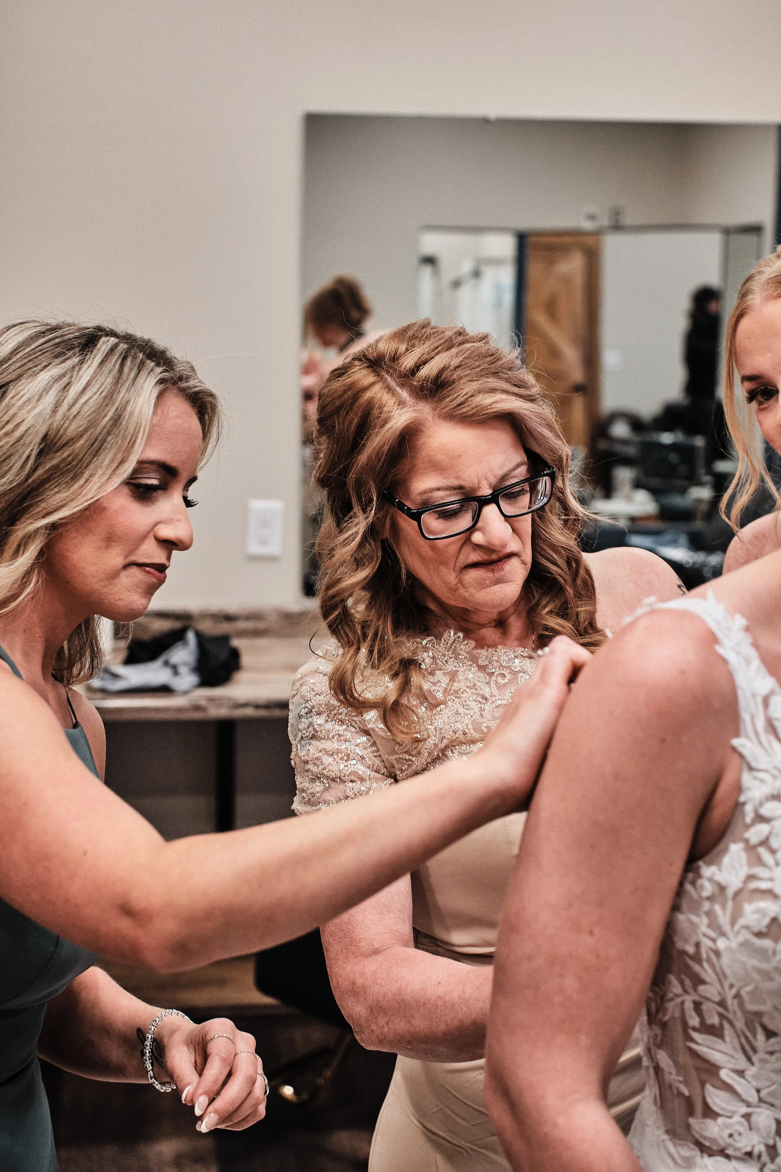 Three women, one wearing glasses, are helping a bride with her wedding dress or veil in a room with a mirror in the background.
