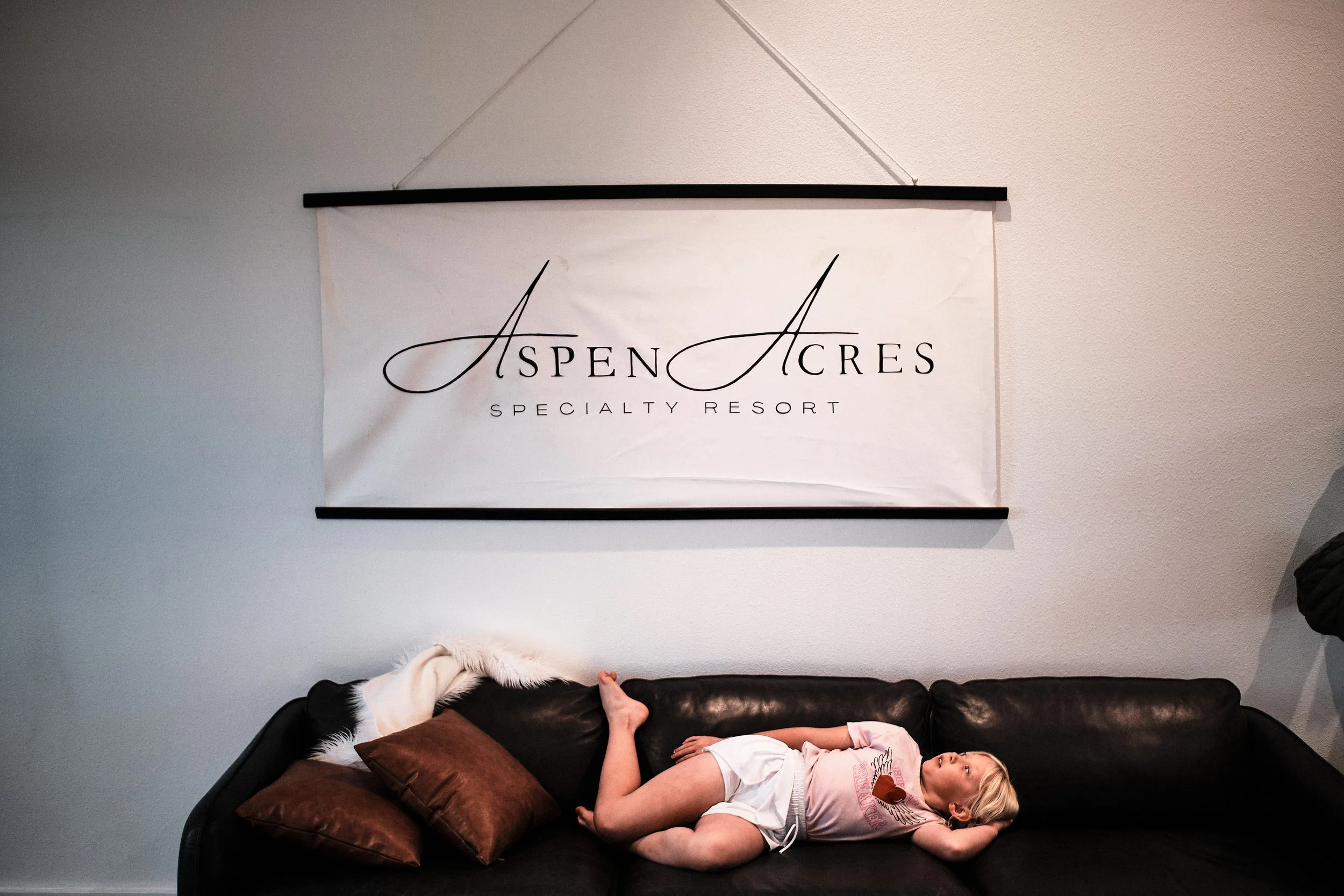 A girl lying on a black leather couch with brown pillows, in front of a wall with a large white sign that reads "Aspen Acres Specialty Resort."