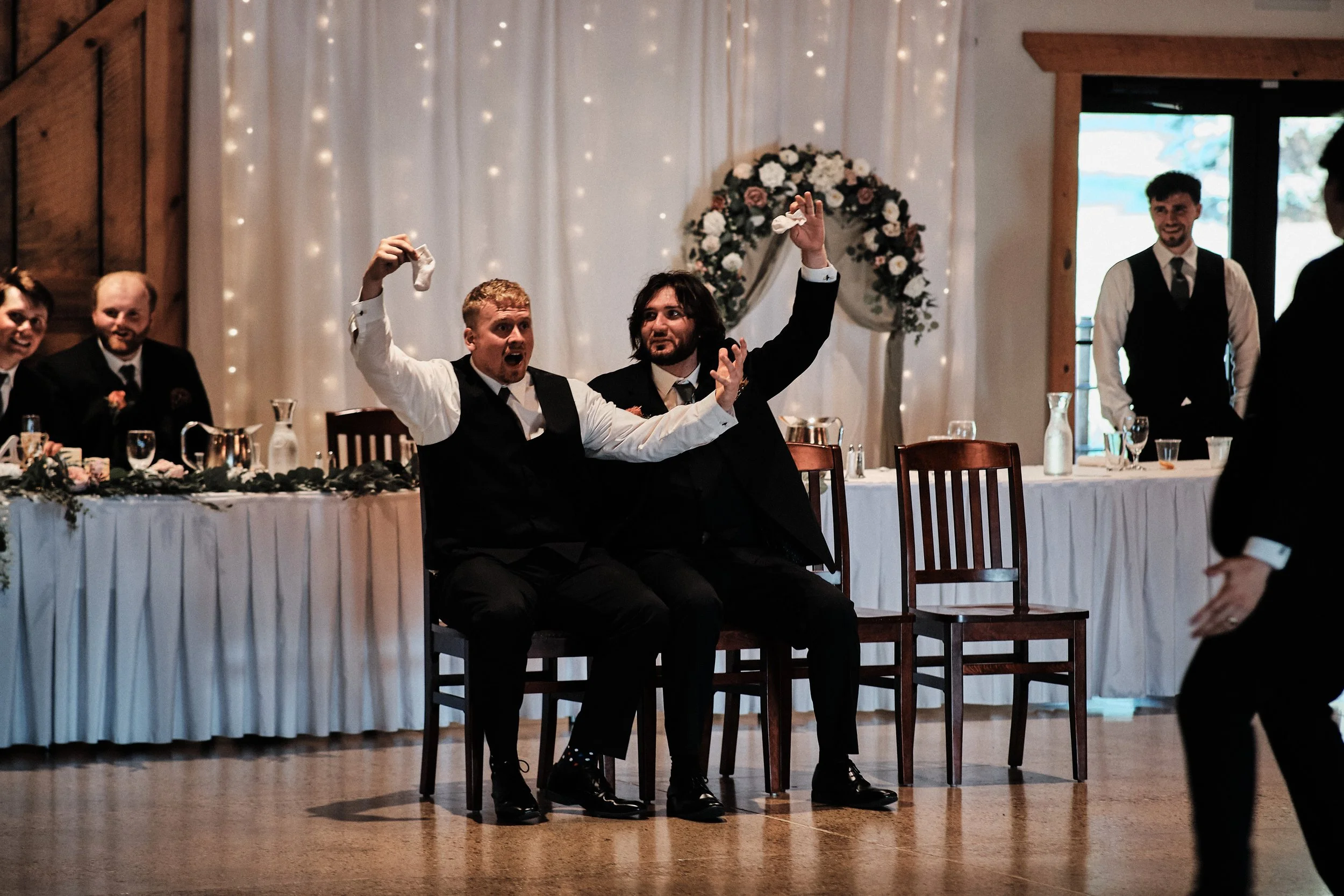 Two men in black suits and vests sitting on chairs at a reception table, raising their hands in a celebratory gesture, with a floral arch and fairy lights in the background and other guests nearby.