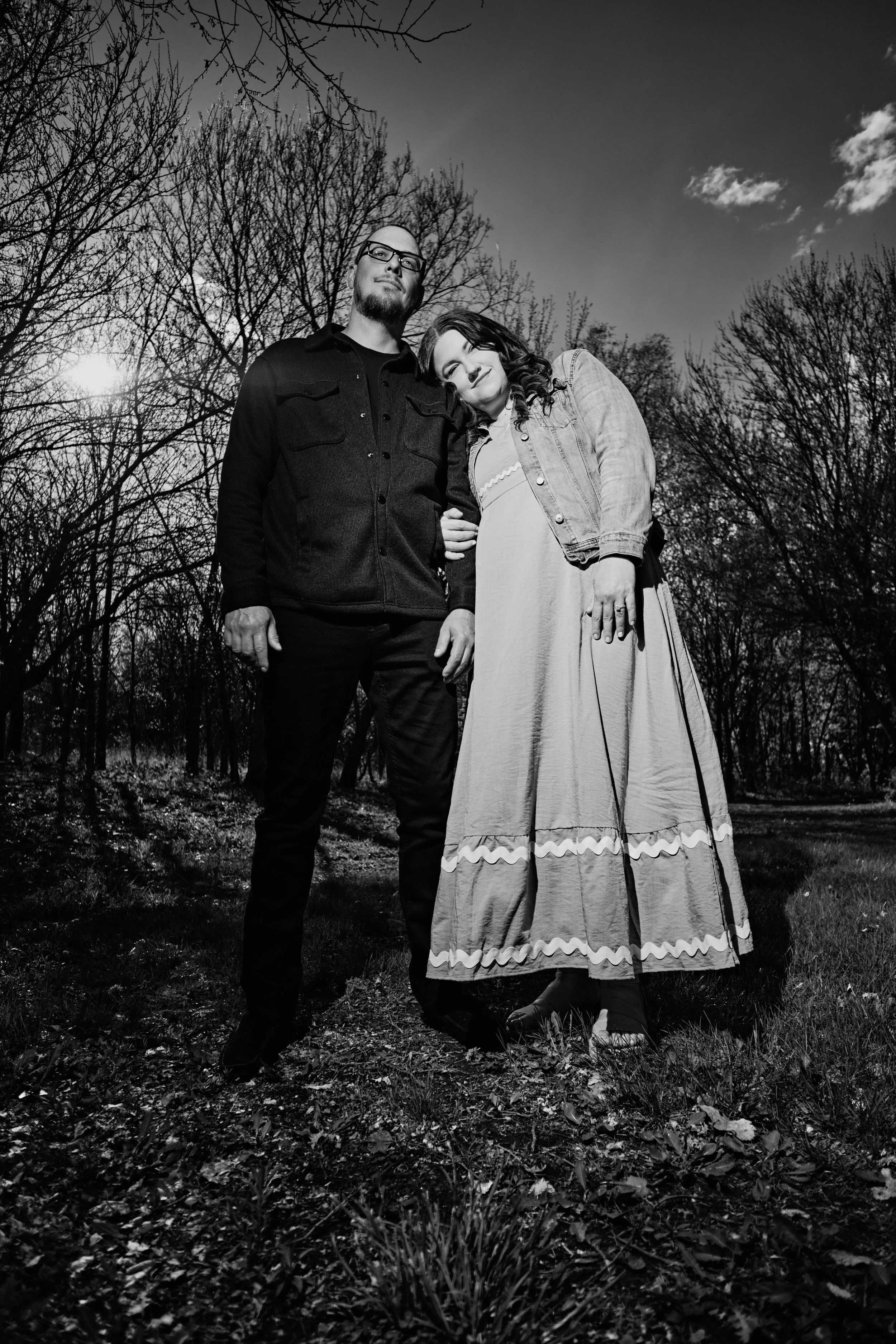 A black and white photo of a couple standing outdoors among leafless trees, with the sun shining partially behind the trees. The man is wearing glasses and a dark jacket, and the woman is leaning her head on his shoulder, wearing a long dress and a d