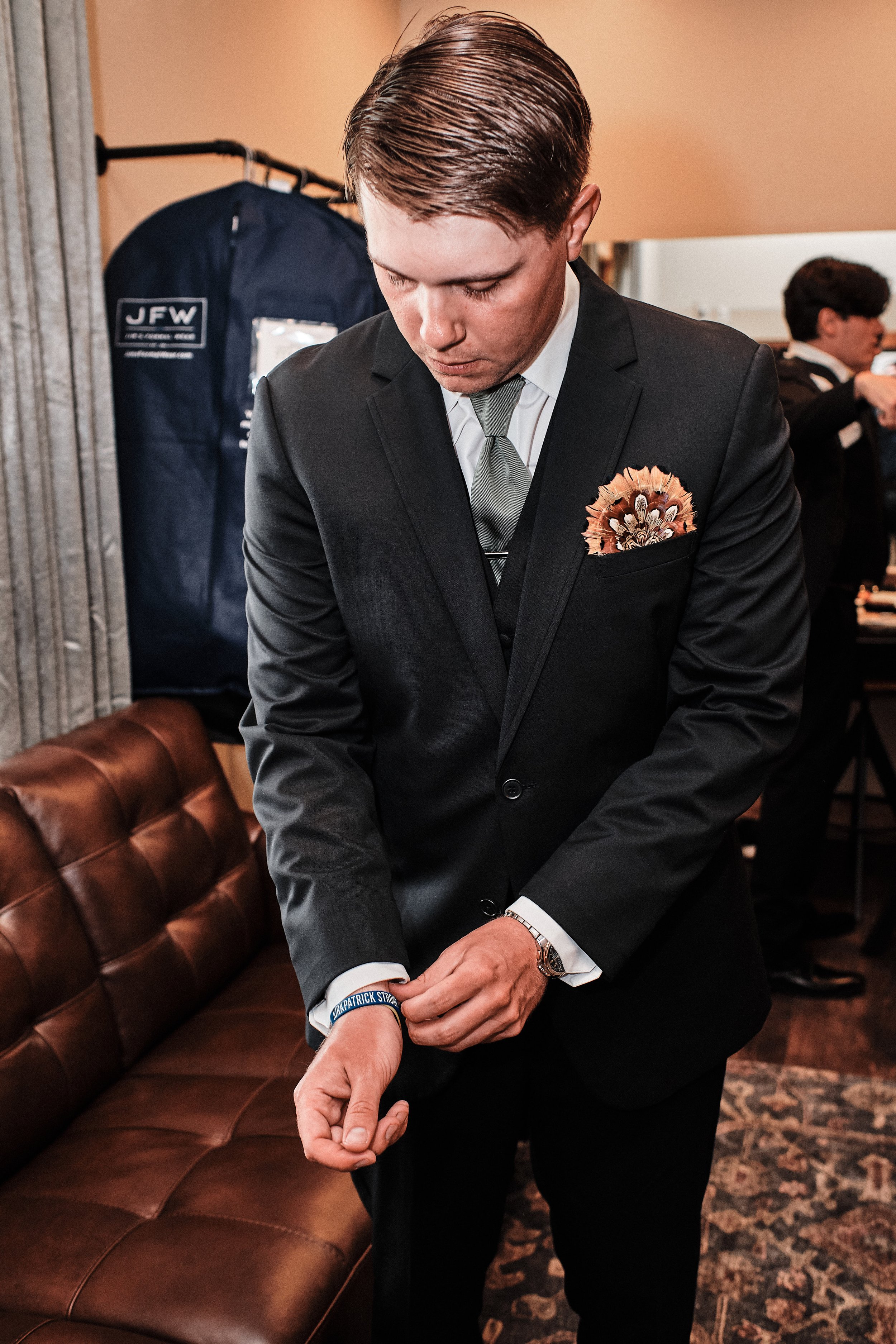 Man in a black suit adjusting his sleeve in an indoor setting with a leather couch and another person in the background.