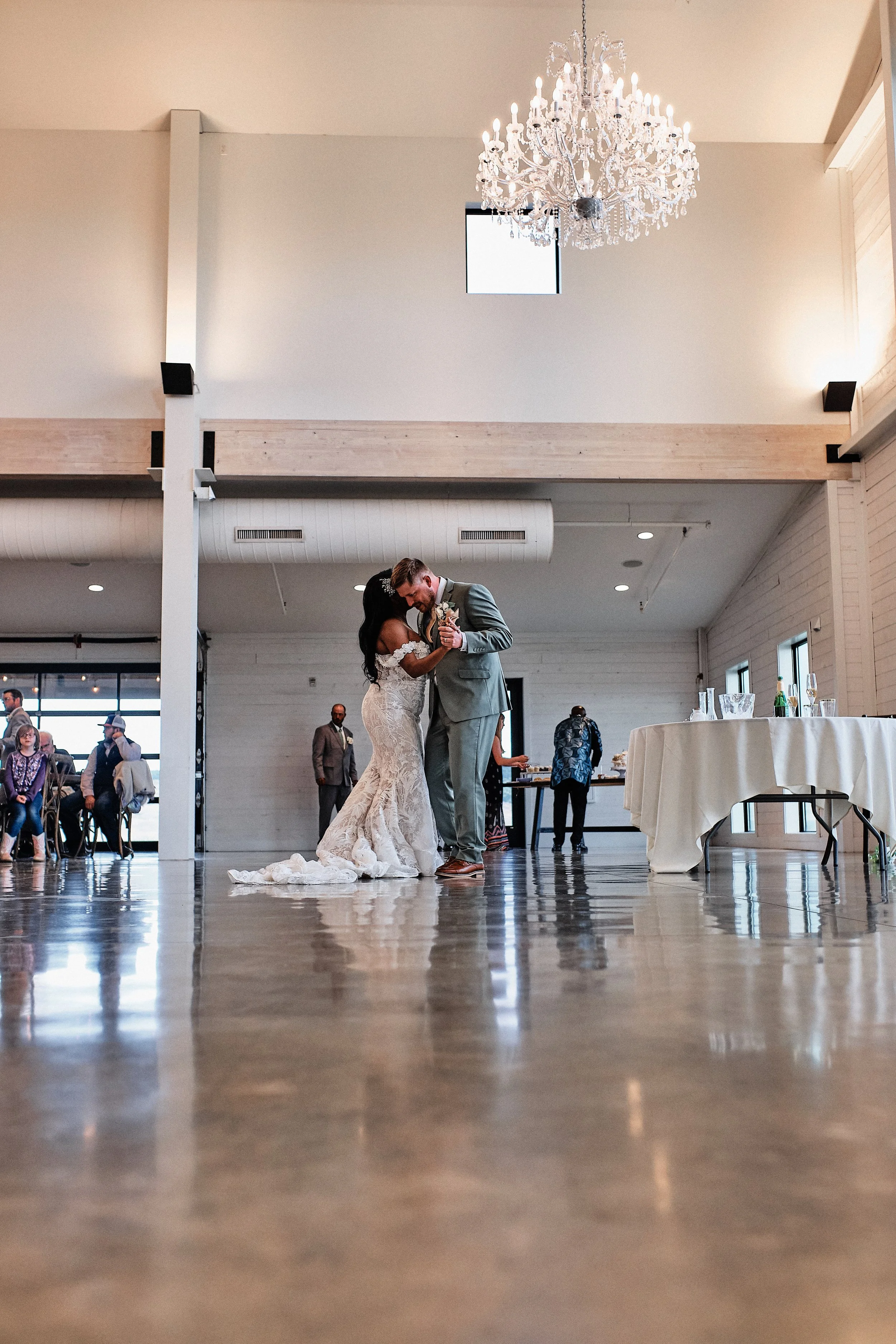 A bride and groom dance closely at their wedding reception in a spacious, well-lit venue with a high ceiling, large windows, and a chandelier. Guests watch from seated positions and stand around the perimeter.