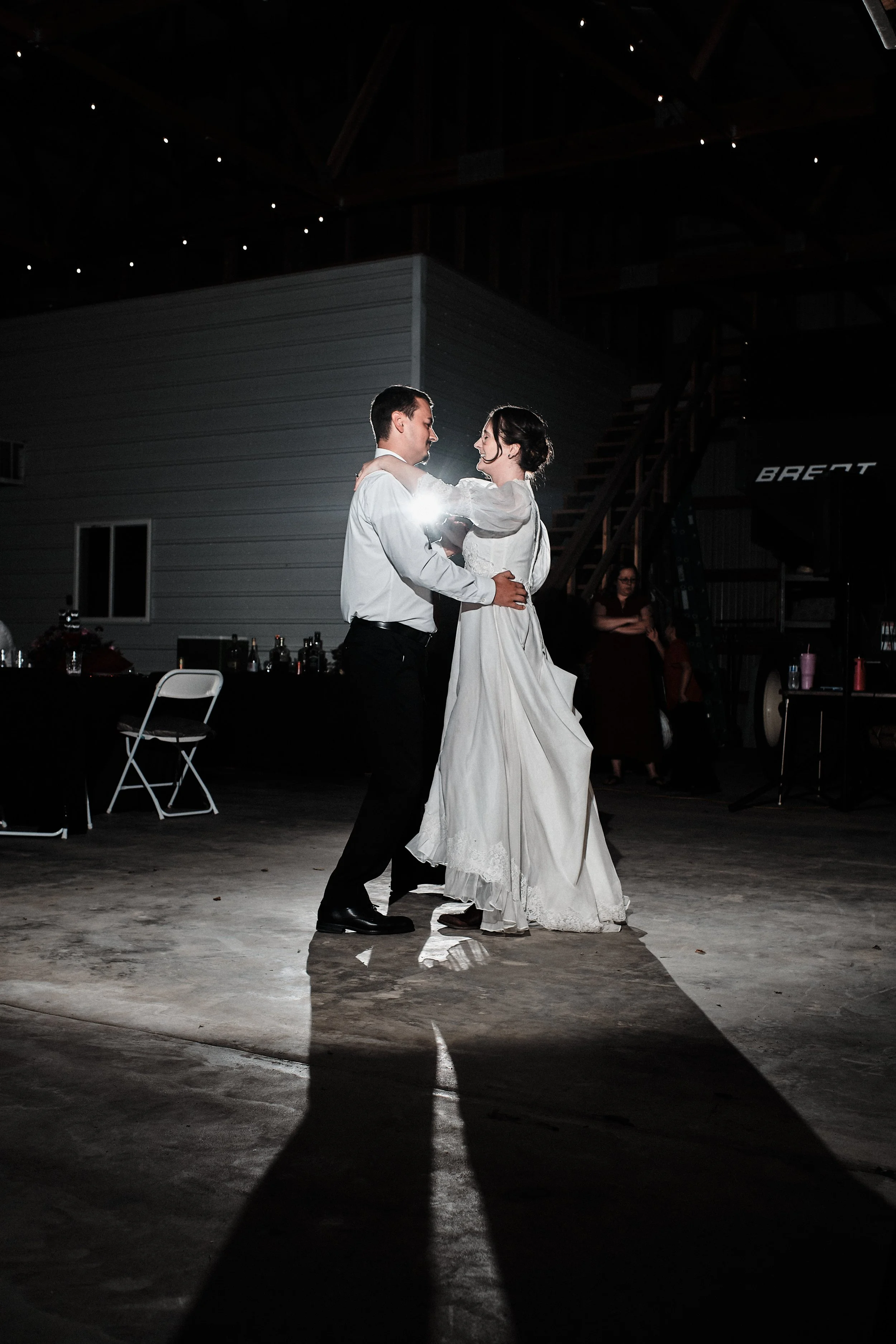 A couple dancing at their wedding reception with a dark background and spotlight highlighting them, with some tables and a person in the background.
