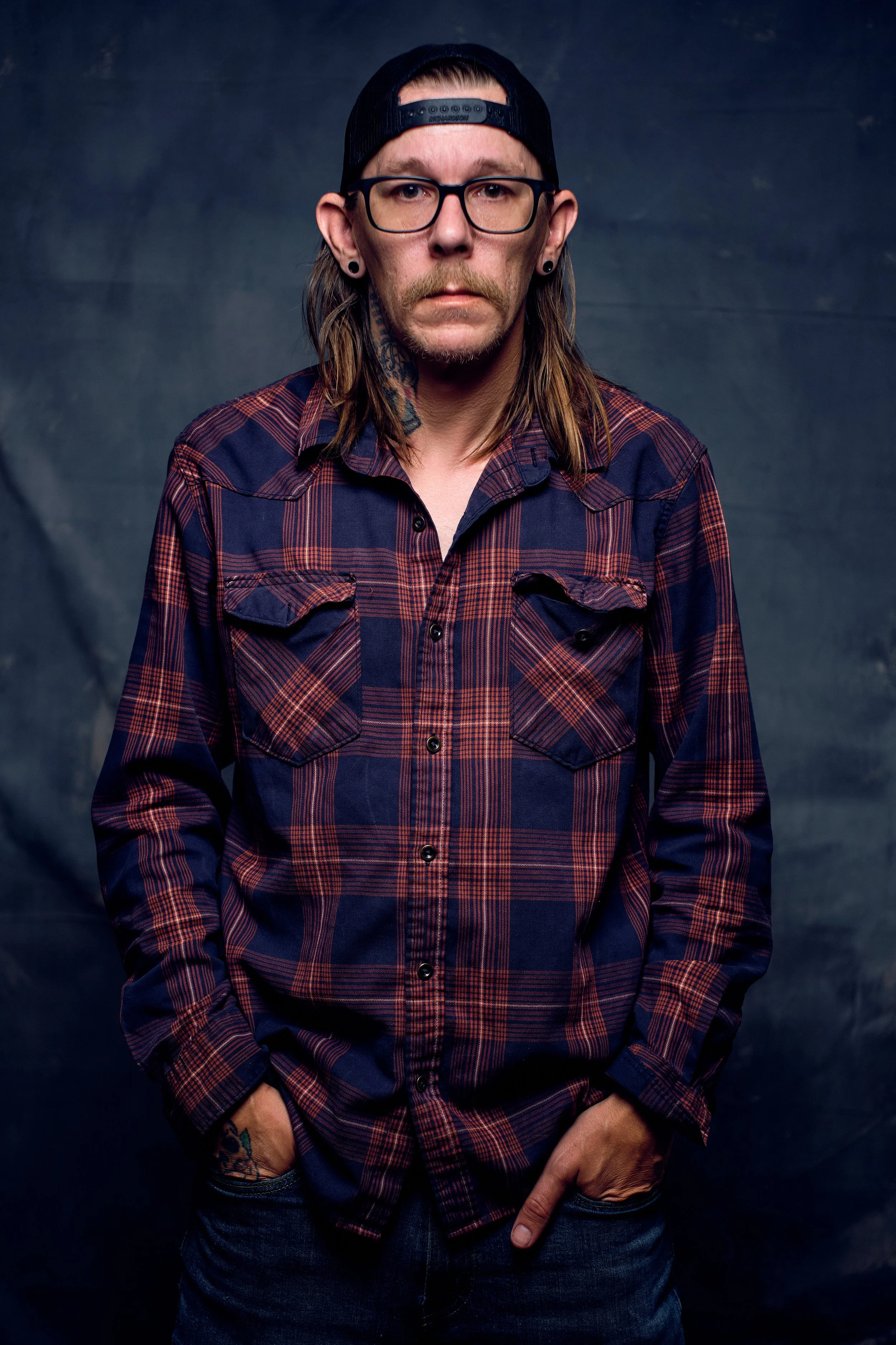 A man with long hair, glasses, tattoos, and facial hair wearing a backwards black cap and a red plaid shirt, standing against a dark backdrop.