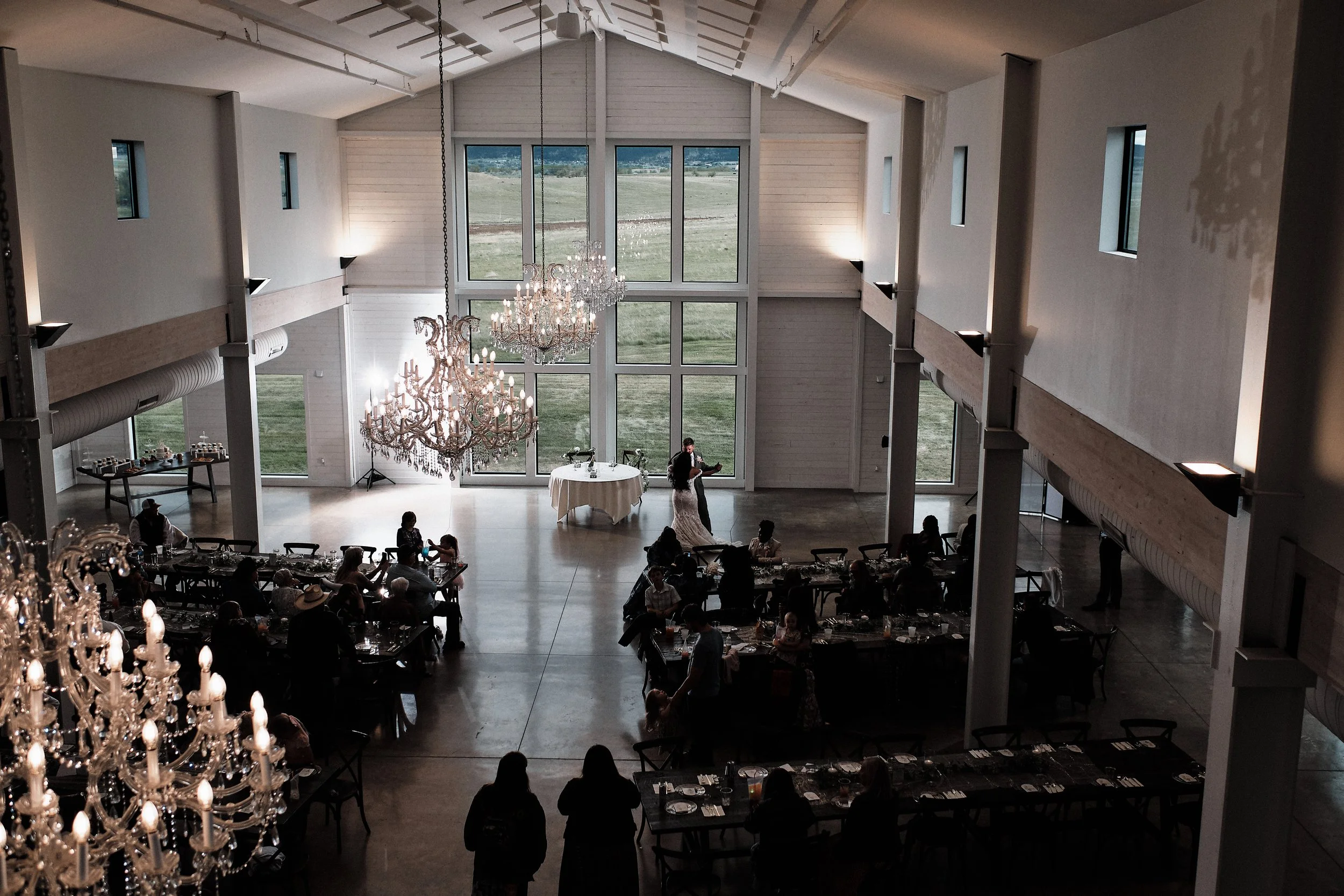 Inside a large, modern wedding venue with a high ceiling, large windows, and elegant chandeliers. A bride and groom are dancing in front of seated guests. The venue has a spacious, rustic interior with white walls and wooden accents.
