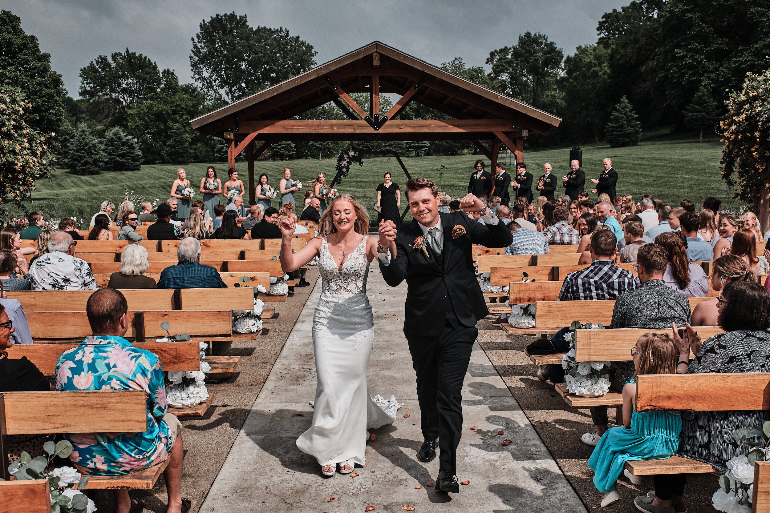 A wedding ceremony outside under a wooden pavilion with a band of bridesmaids and groomsmen. The bride and groom are walking down the aisle, holding hands and smiling. Guests are seated on wooden benches, and the background features lush green trees 