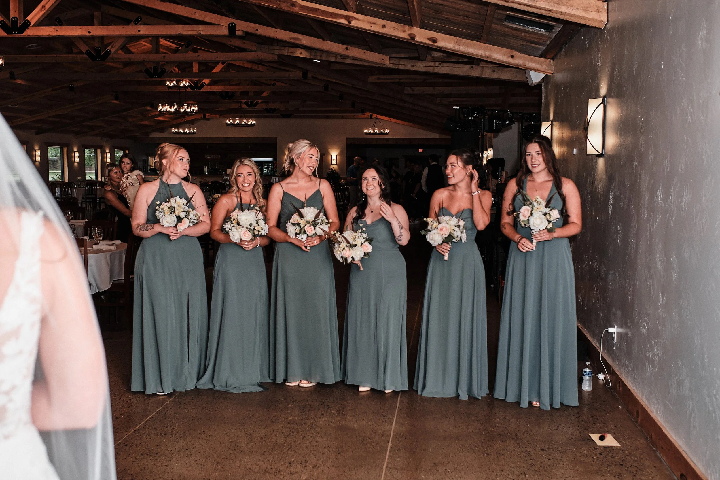 A group of six women in matching teal bridesmaid dresses holding bouquets, standing in a reception hall with wooden vaulted ceilings and dim lighting, smiling and talking.