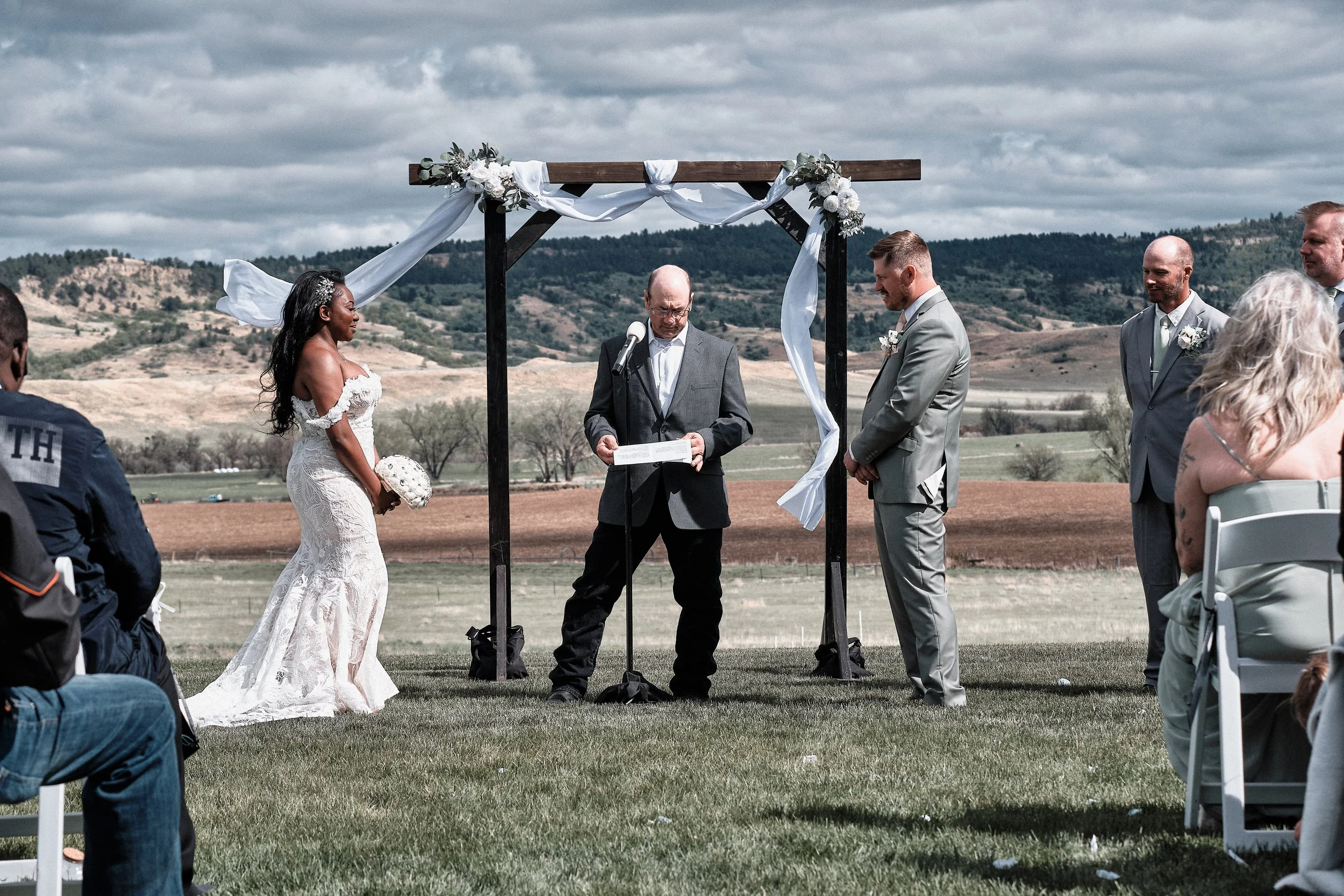 A couple getting married outdoors, holding hands and facing each other during the ceremony, with a scenic backdrop of hills and sky, under a wooden arch decorated with white fabric and flowers.