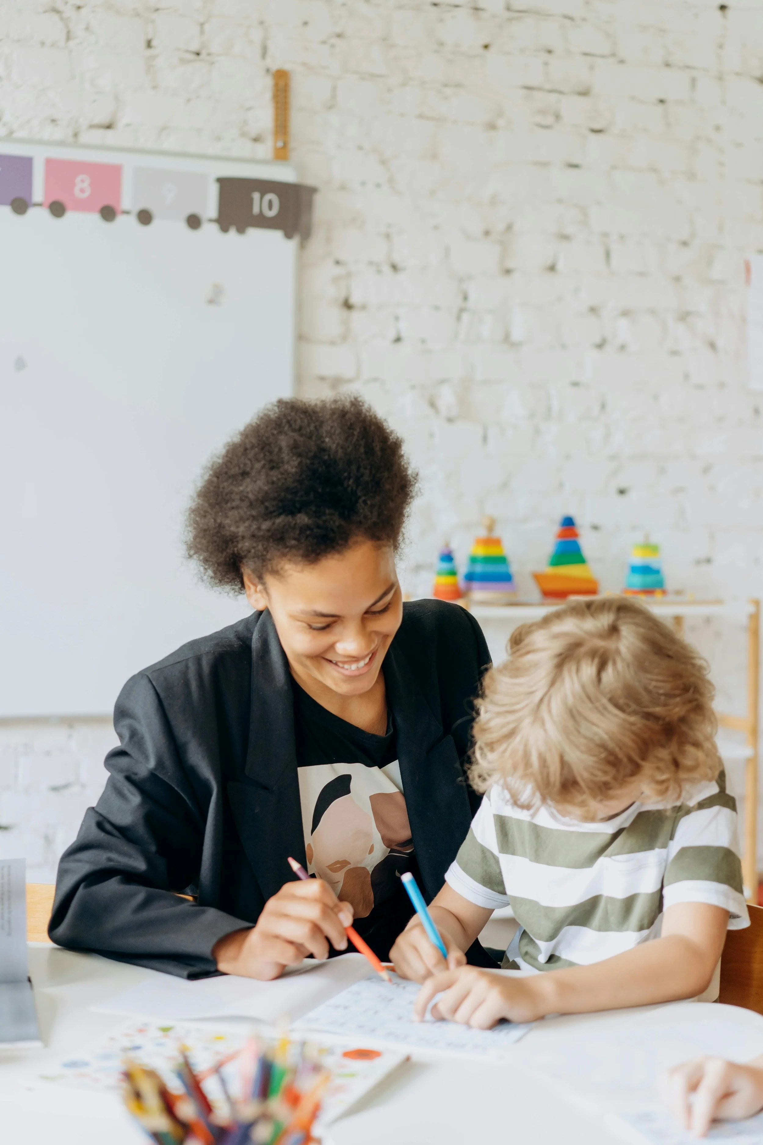 A woman and a young boy are sitting at a table, writing and drawing with colored pens. The woman is smiling and looking at the boy. The background features a white brick wall, educational decorations including a colorful train and stacking rainbow cones, and art supplies on the table.