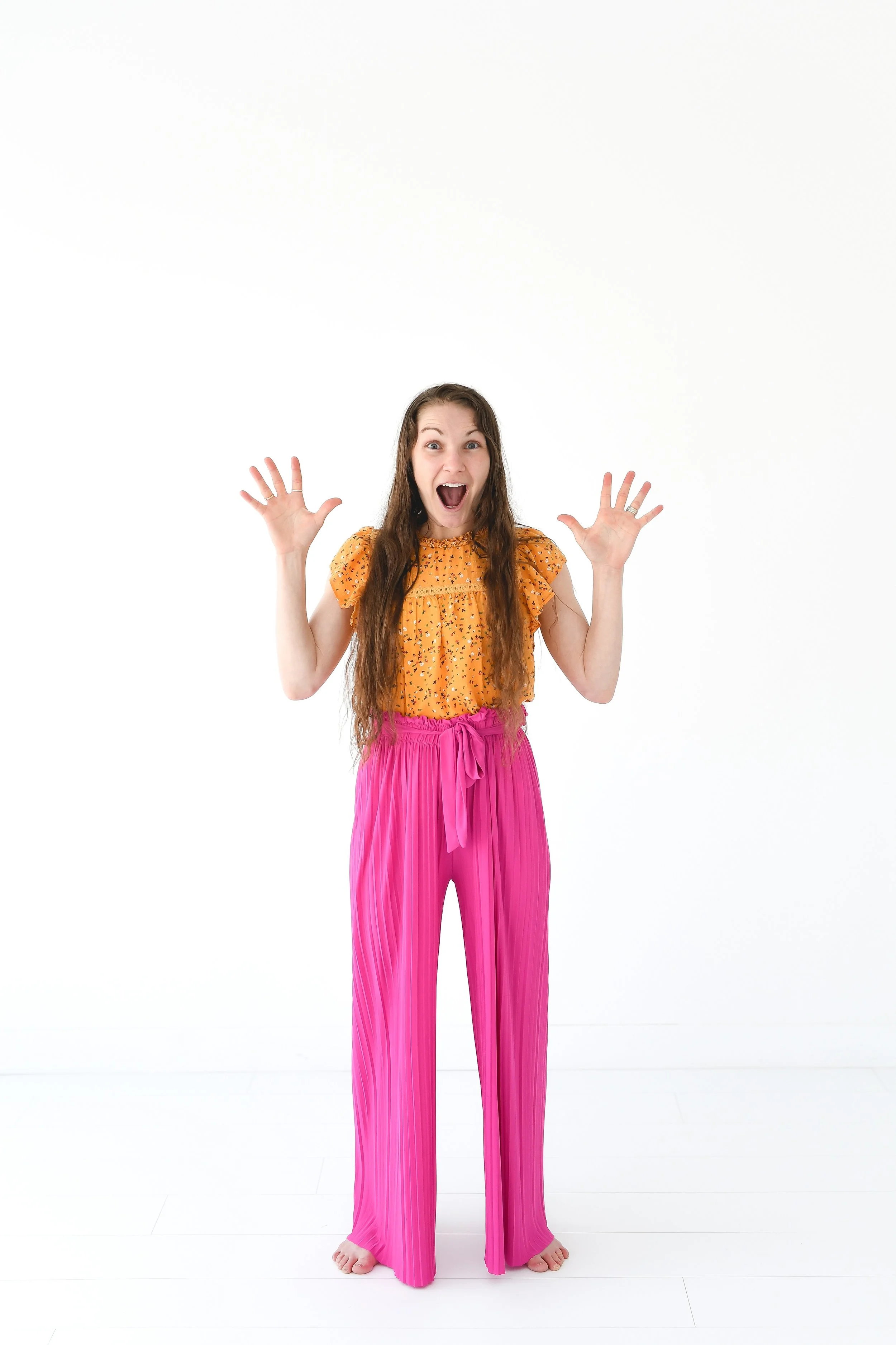 A woman with long, wavy brown hair is standing barefoot with her feet apart against a plain white background. She is wearing a sleeveless orange top with a floral pattern and bright pink wide-leg pants with pleats, tied at the waist. She has an excited expression with her mouth open and hands raised near her head, fingers spread apart.