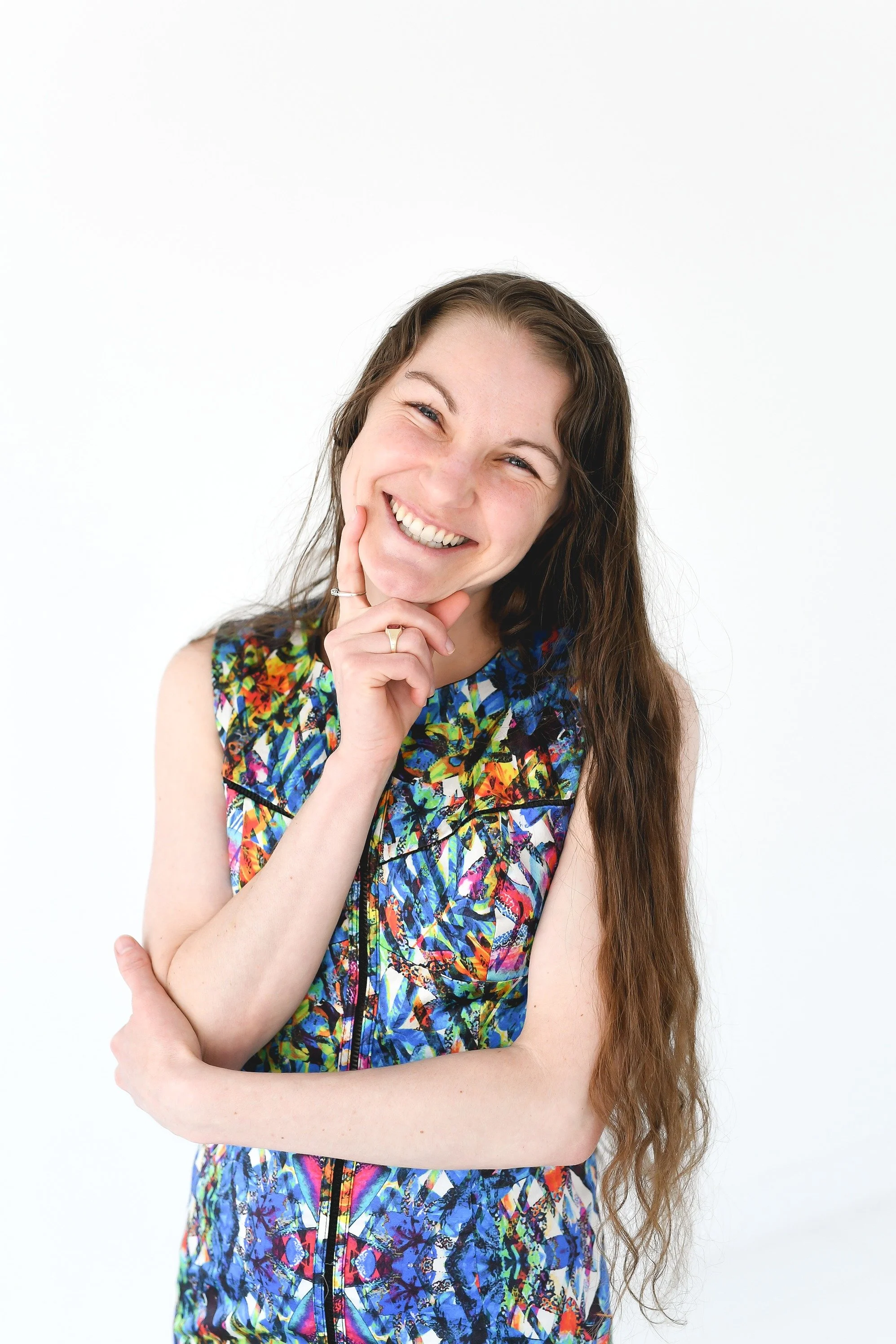 Smiling woman with long, wavy brown hair wearing a colorful, patterned sleeveless dress, posing with her hand on her chin against a plain white background.