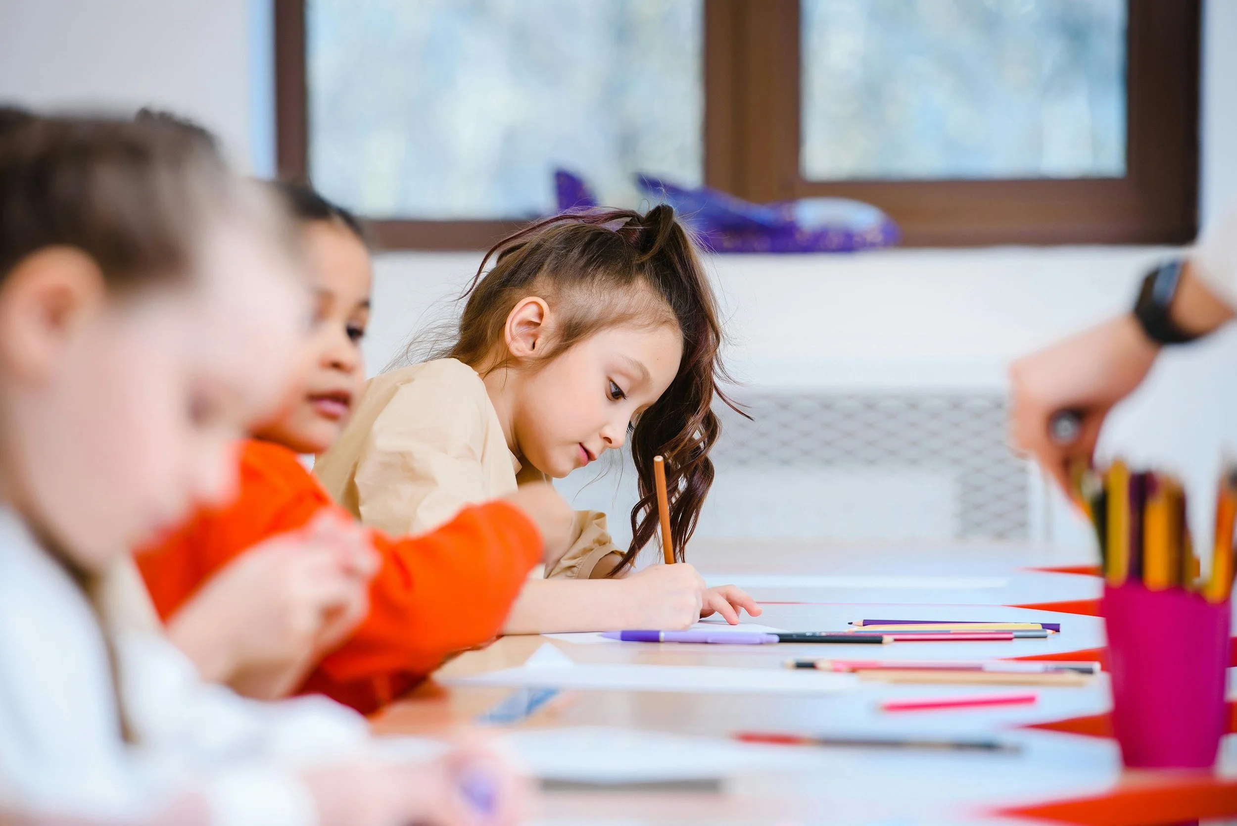 Children sitting at a classroom table, writing with pencils, with colorful supplies in front of them, and a window in the background.