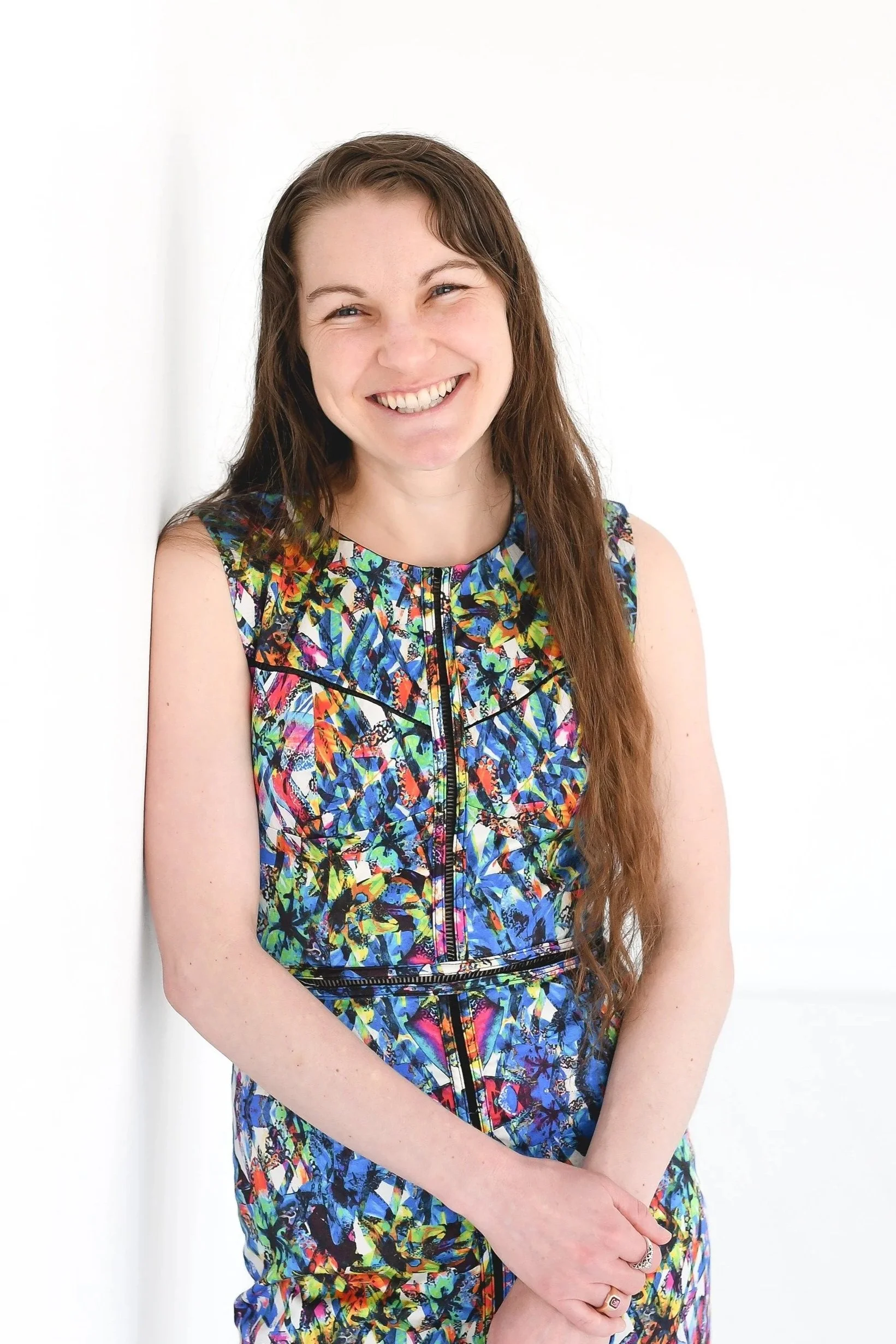 A young woman with long brown hair smiling, wearing a colorful abstract-patterned dress, standing against a plain white background.