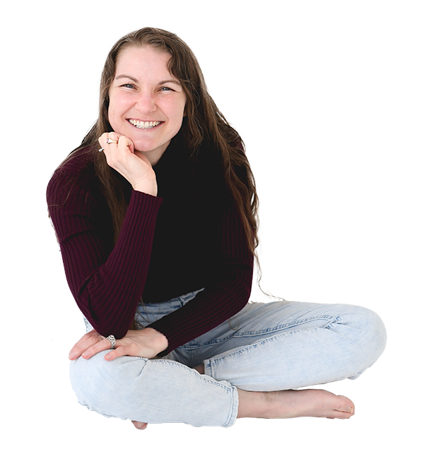 A young woman with long brown hair, smiling, sitting cross-legged on the floor, wearing a dark red/purple sweater and light blue jeans.