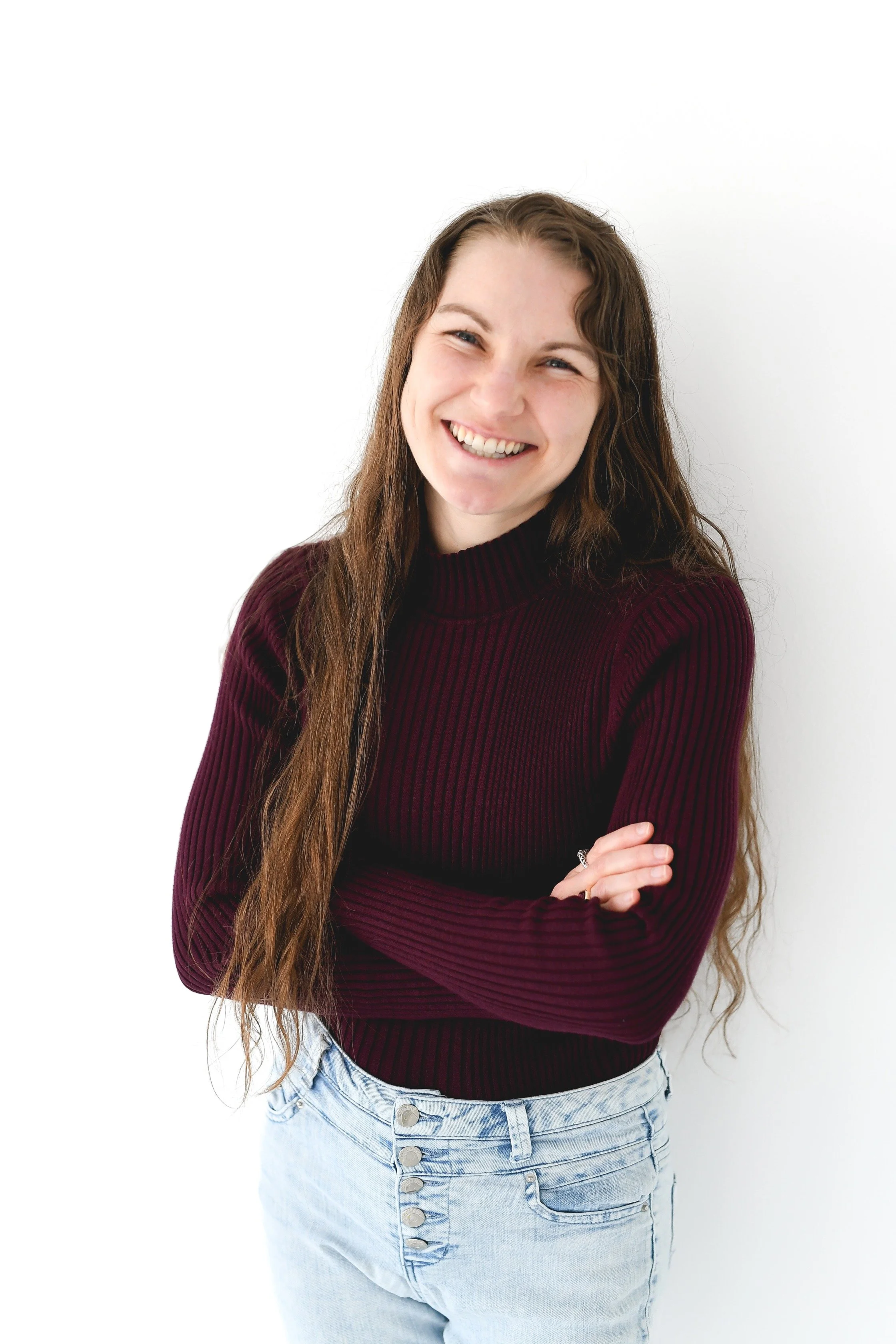 Young woman smiling with long brown hair, wearing a maroon sweater and light blue jeans against a white background.