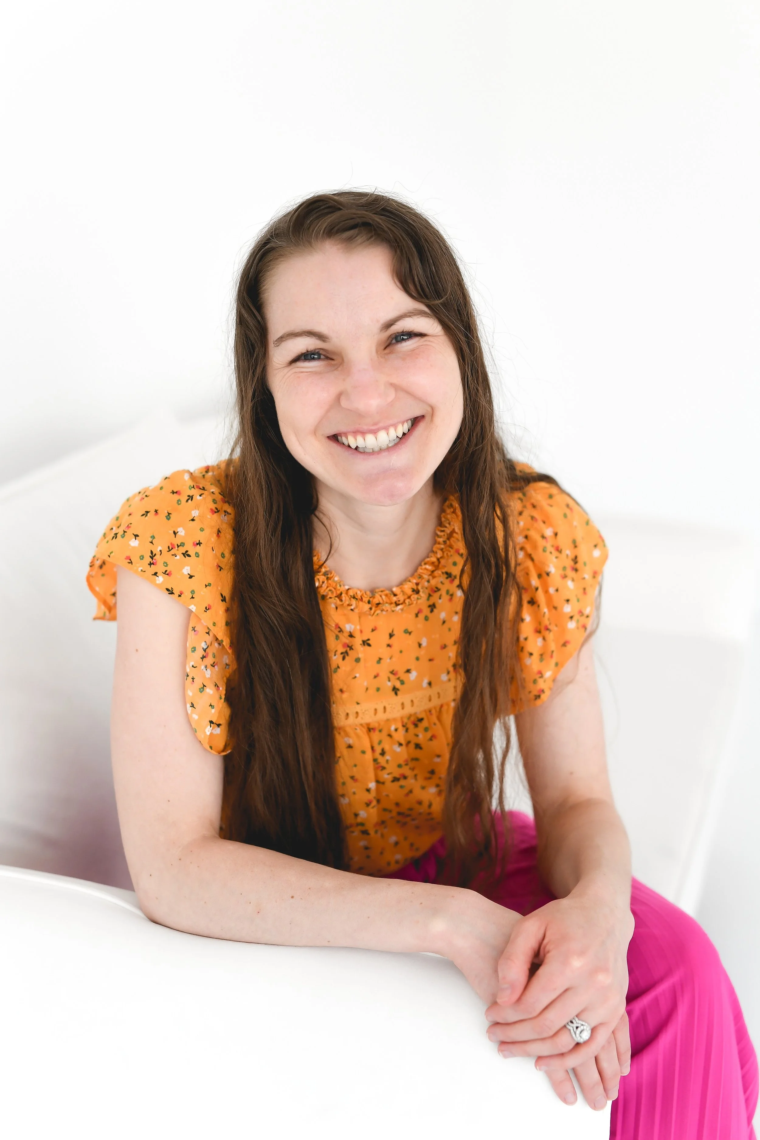A woman with long brown hair and blue eyes, smiling, sitting at a white table, wearing a yellow floral top and pink pants, against a white background.