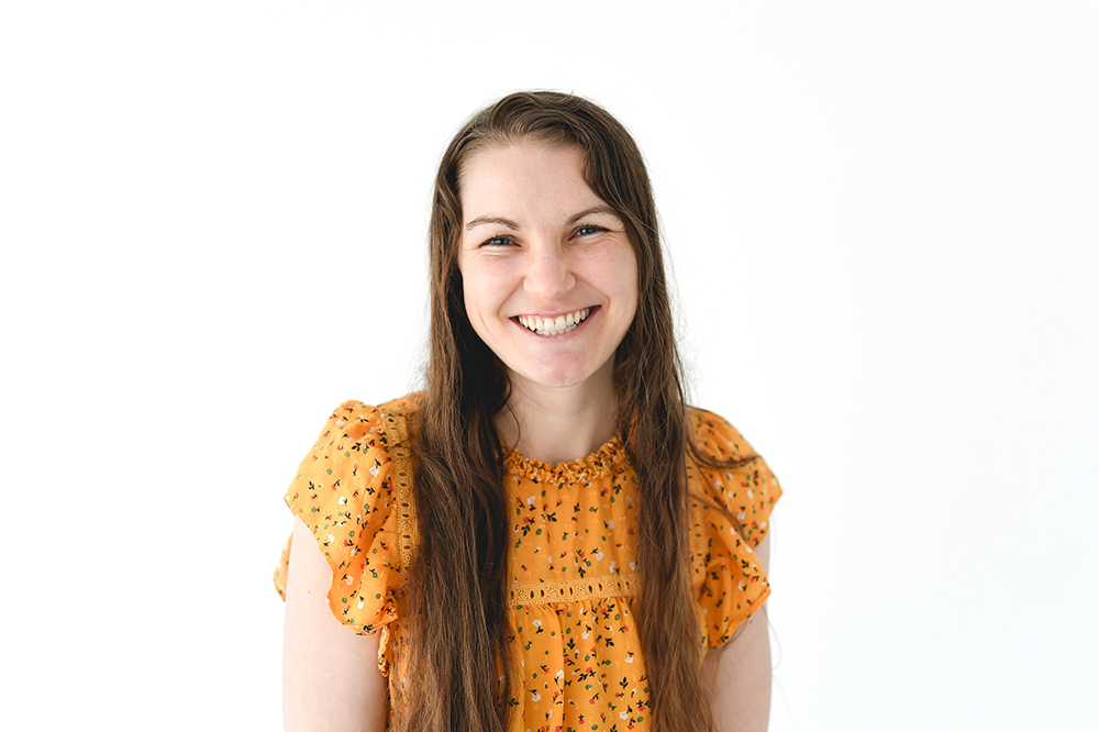 Young woman with long brown hair wearing an orange floral dress smiling against a white background.