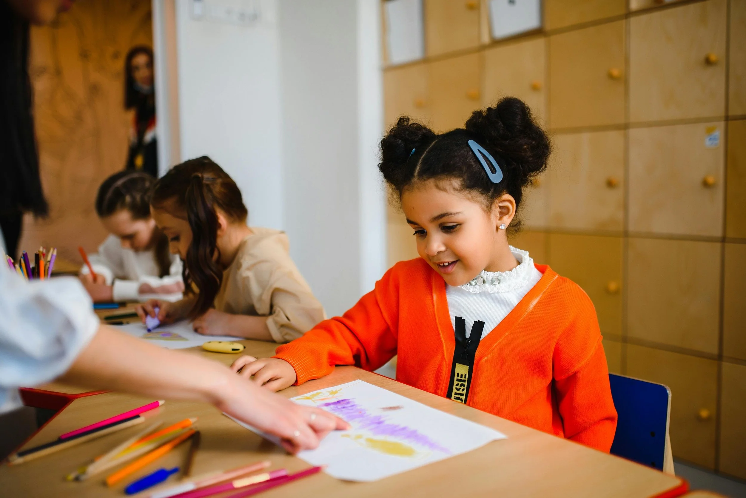 Young girl with curly hair styled in buns, wearing orange jacket and earrings, smiling while engaging in drawing or coloring activity at a table in a classroom. Other children are also focused on their art projects, and art supplies like colored pencils are scattered on the table.