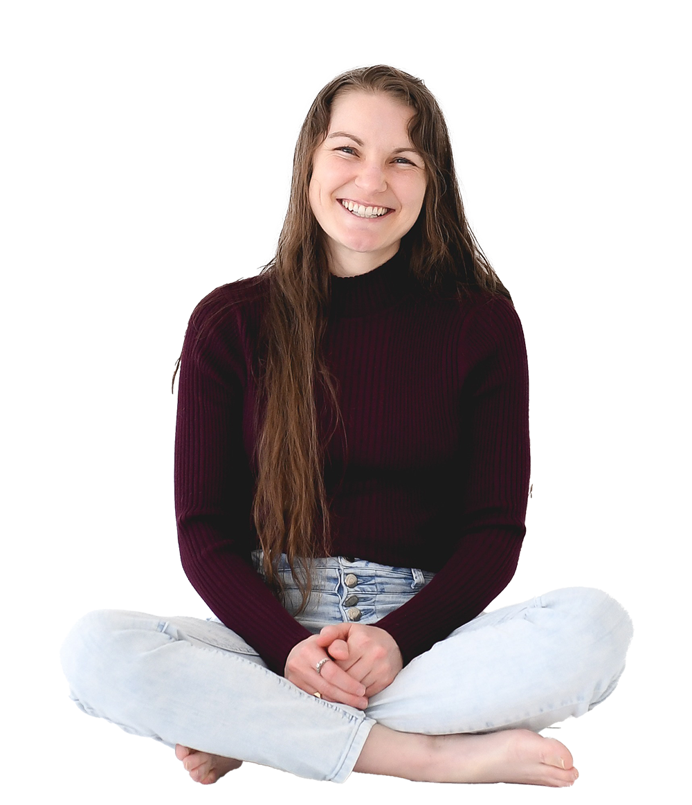A young woman with long brown hair sitting cross-legged on the floor, smiling, wearing a dark red sweater and light blue jeans.