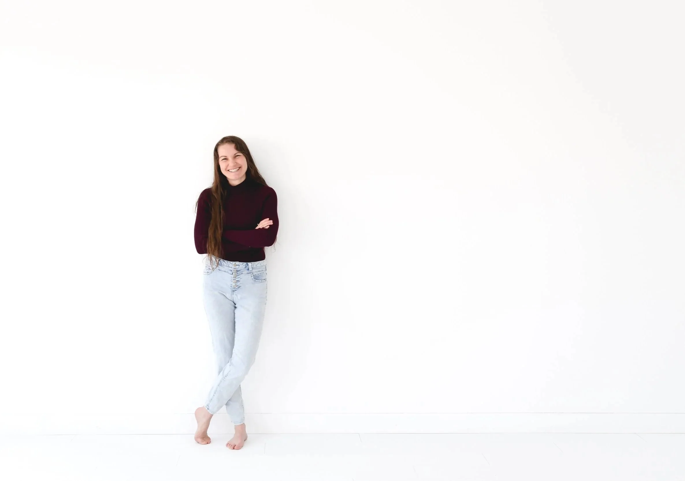 A young woman with long brown hair, wearing a dark burgundy sweater and light blue ripped jeans, smiling with arms crossed, standing with one foot slightly in front of the other against a white background.