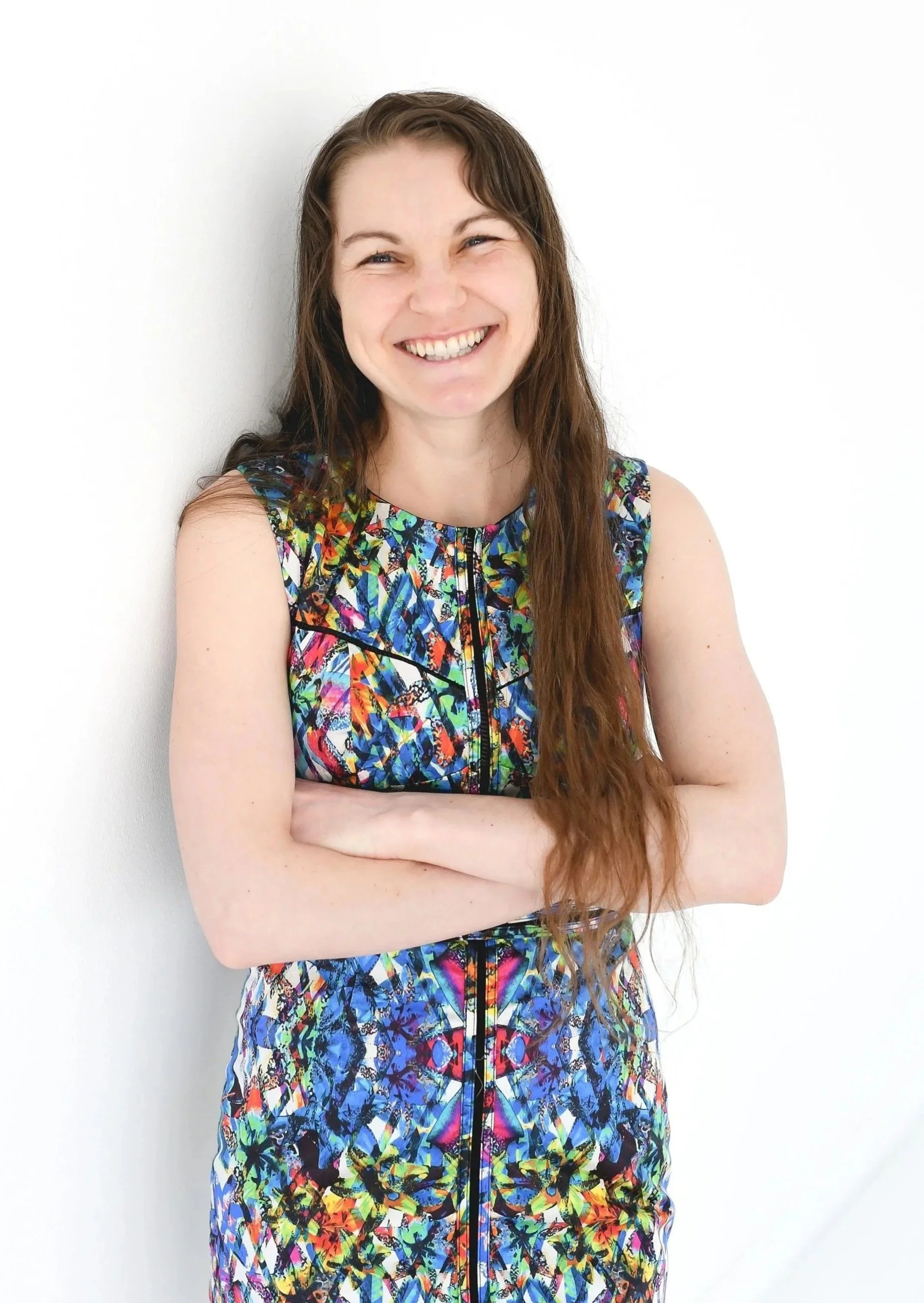 Portrait of a smiling woman with long wavy brown hair, wearing a sleeveless, colorful, abstract pattern dress, arms crossed, standing against a plain white background.