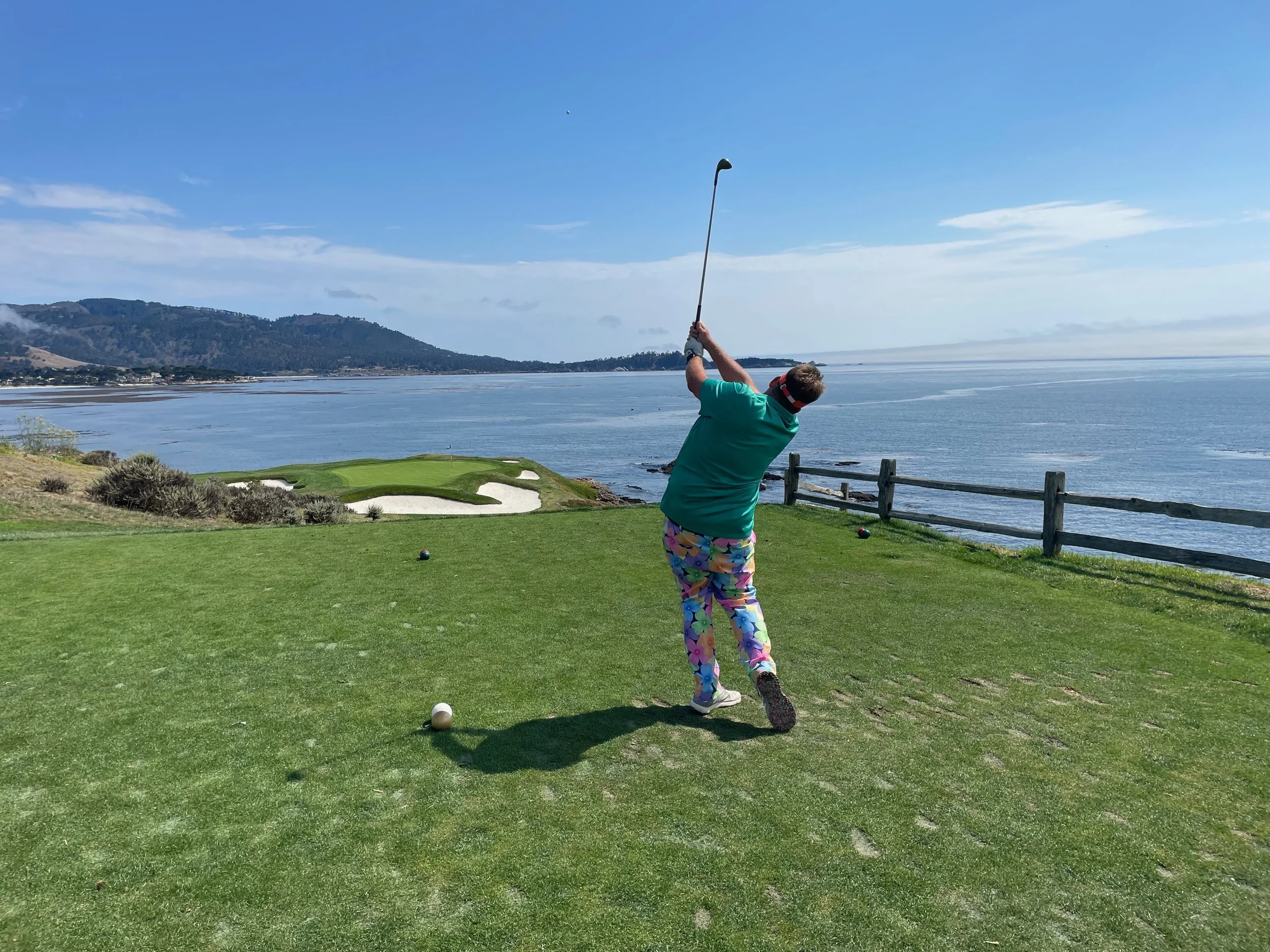 Founder and COO Rob Doten in colorful shorts and teal shirt swinging a golf club on Pebble Beach golf course near the ocean, with hills in the background.