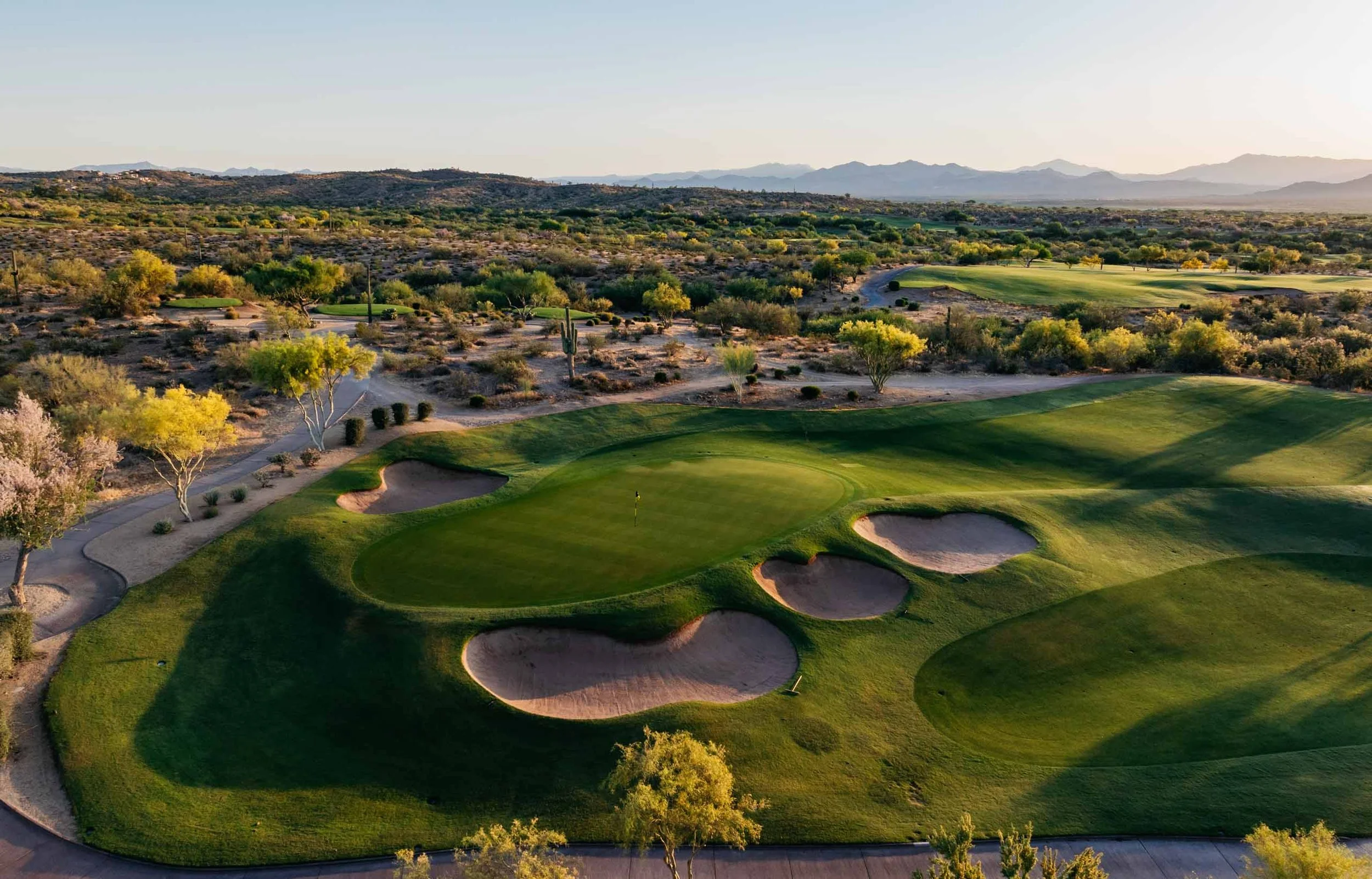 An aerial view of a golf course with lush green fairways and sand bunkers, surrounded by desert terrain with sparse trees, set against distant mountains under a clear sky.  We-Ko-Pa Casino Resort, Arizona, Cholla.